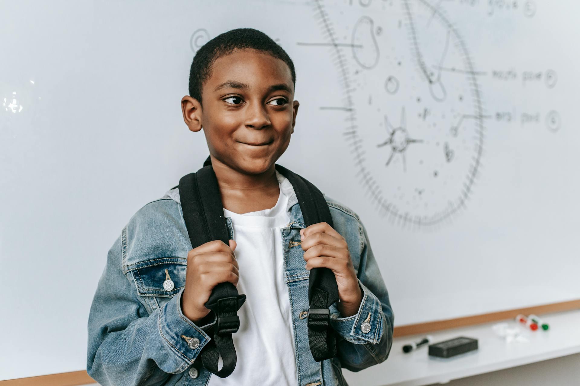 A child wearing a denim jacket and black backpack stands in front of a whiteboard with scientific illustrations.