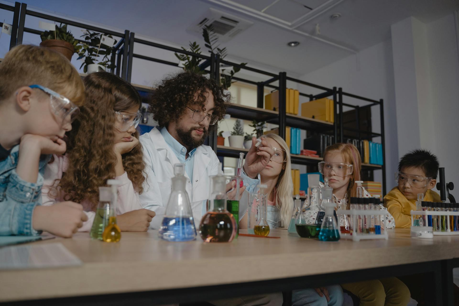 A teacher in a lab coat instructs children seated at a table with various colorful chemistry glassware in a classroom setting.
