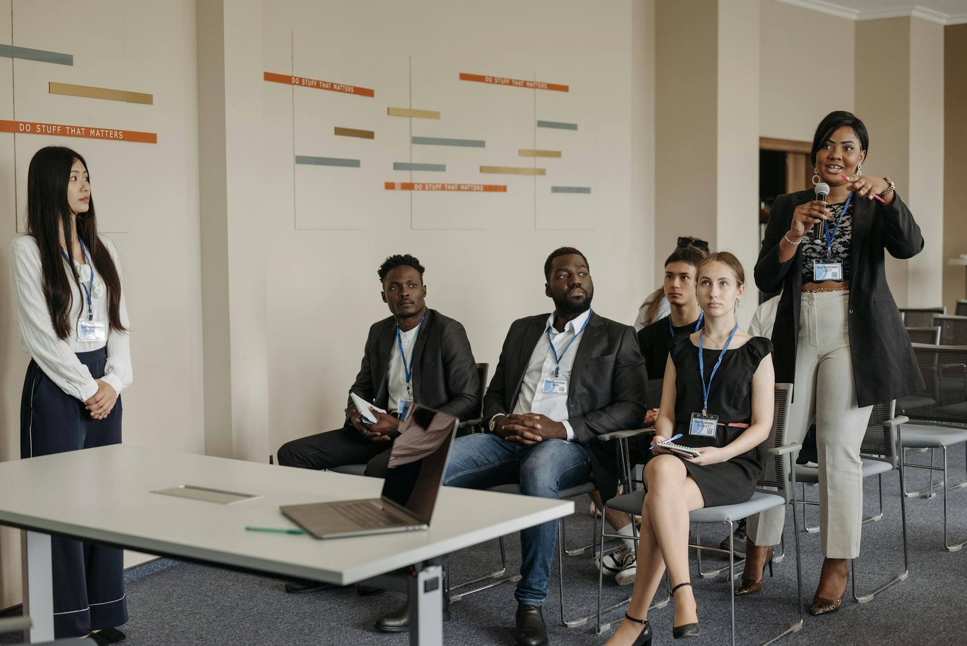 A professional woman stands and speaks with a microphone to a group of seated colleagues in a modern meeting room.