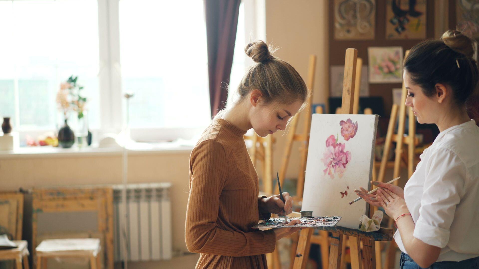Two women paint a floral picture on canvases in a sunlit art studio.
