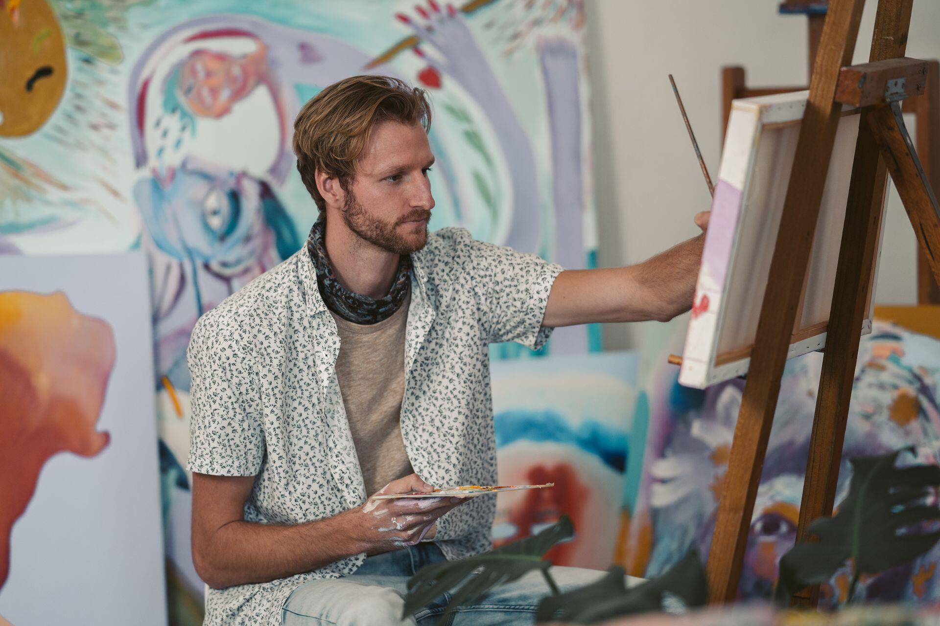 A man with blond hair and a beard paints on a canvas on an easel, surrounded by abstract paintings.