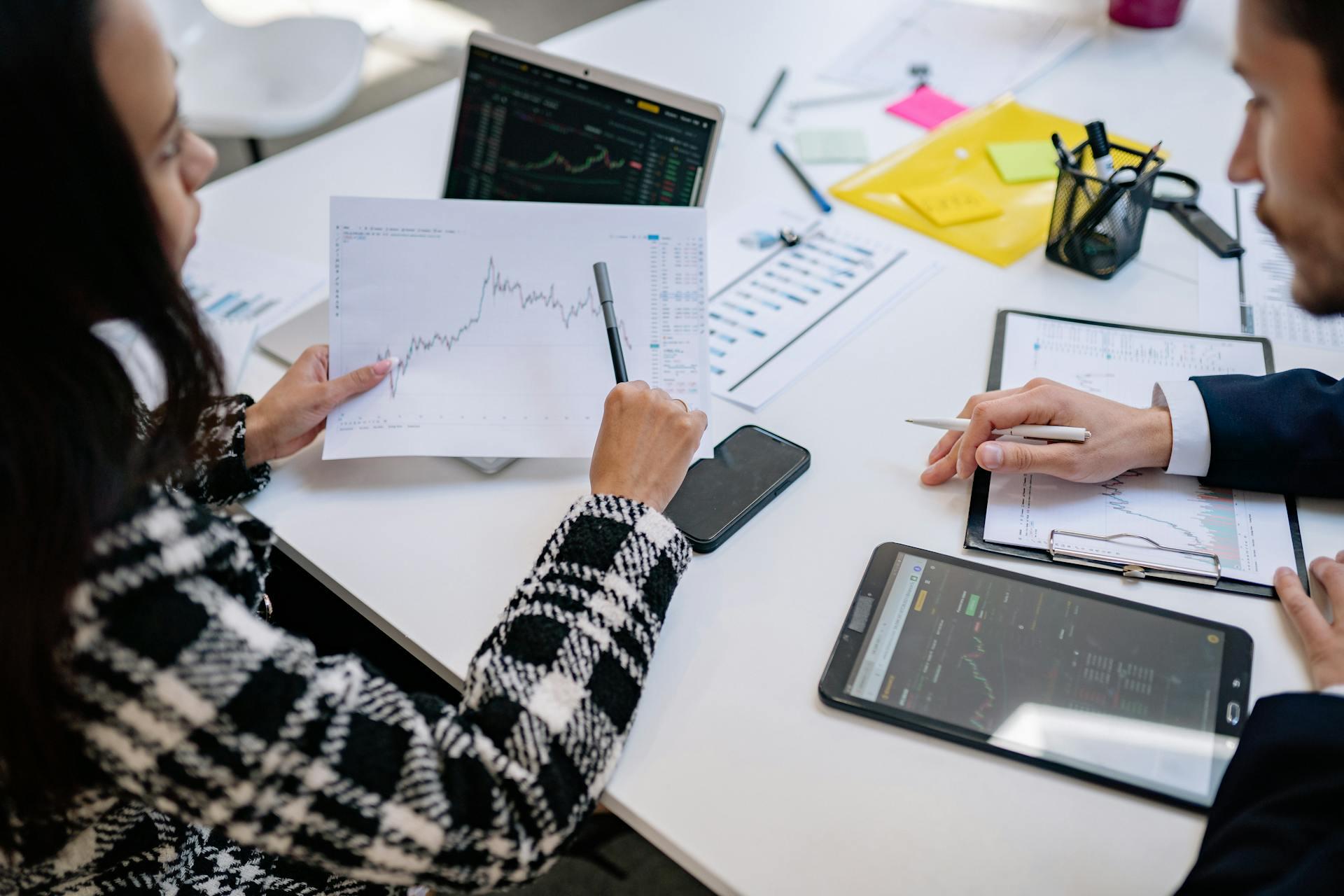 A business meeting featuring hands reviewing market charts and data on laptops and tablets, surrounded by office supplies and documents.