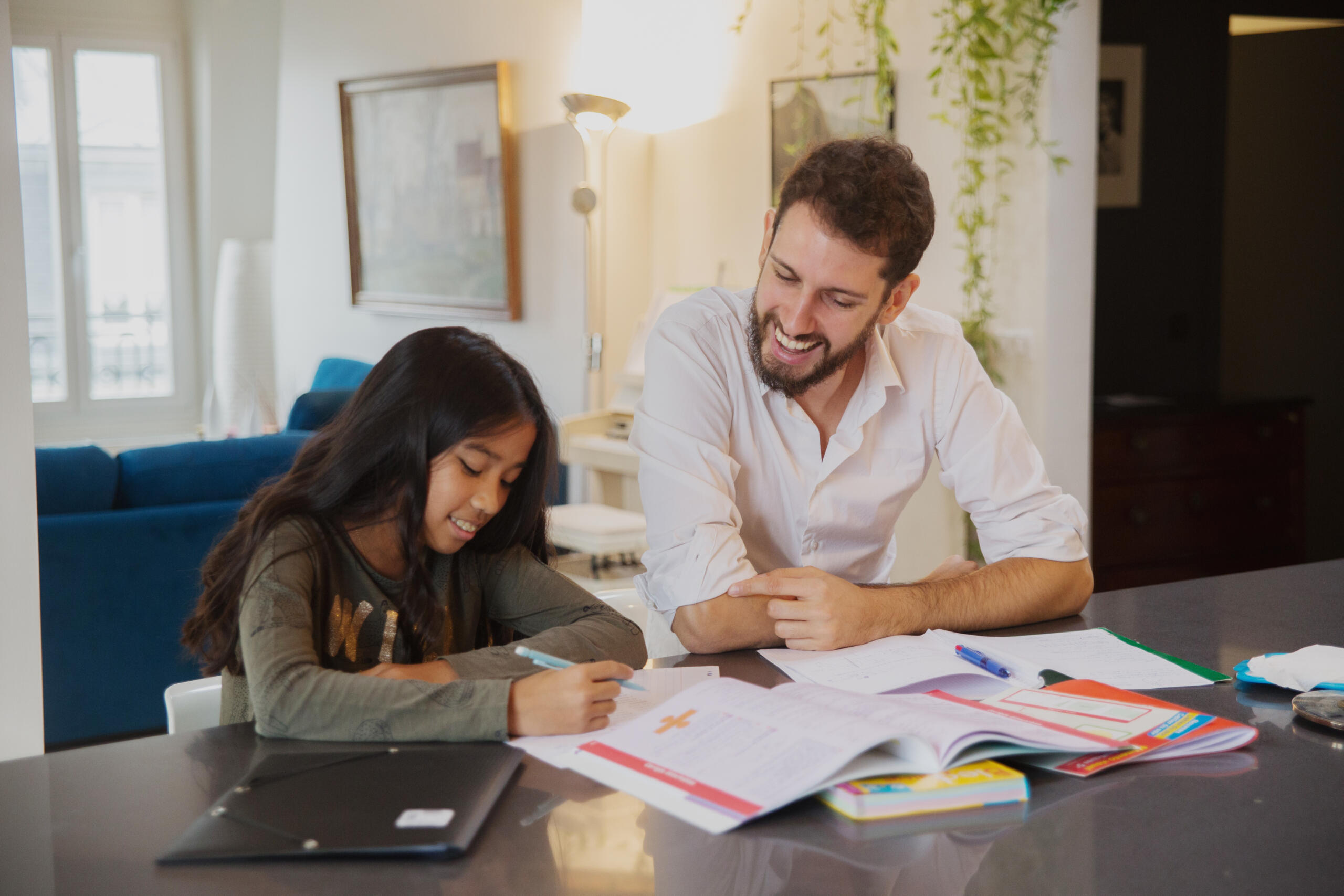 Male tutor helping a young girl with her homework at a kitchen table, both smiling and engaged in the lesson.