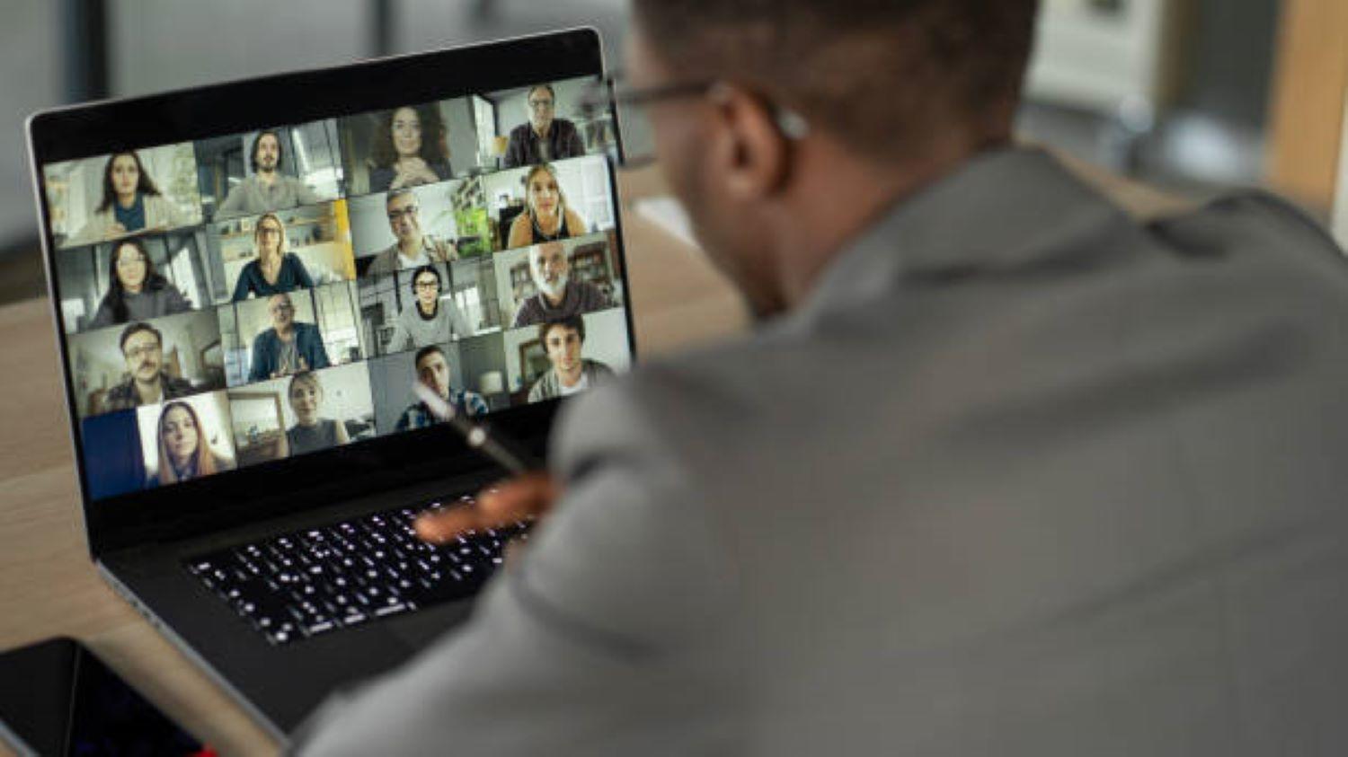 A man looks at a laptop while a virtual meeting happens onscreen.