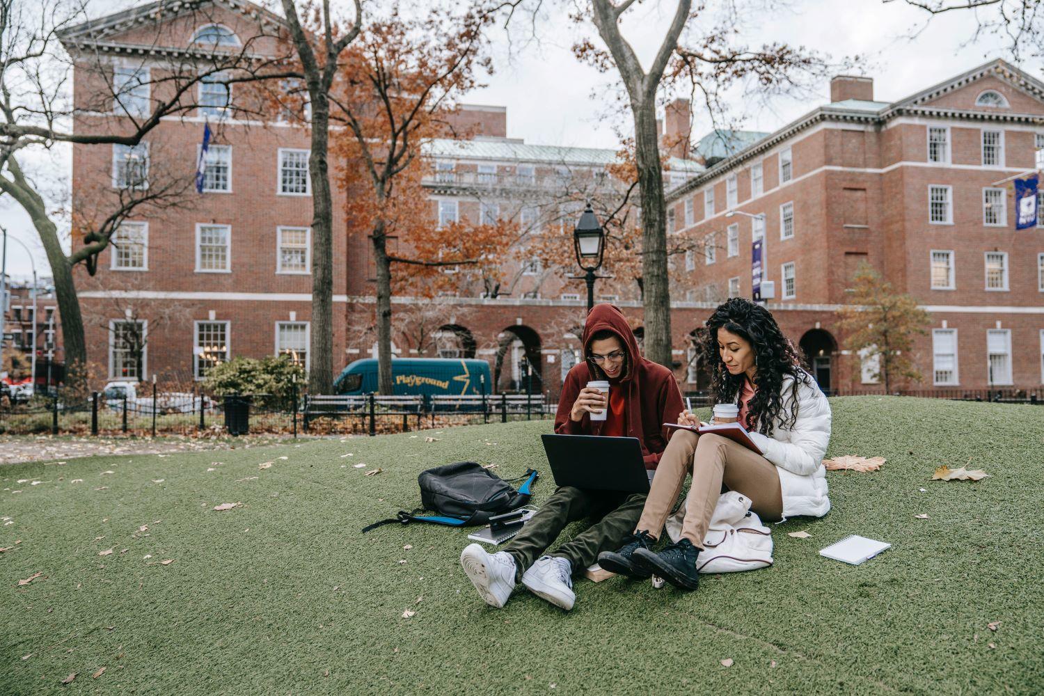 Two college students sit on a grassy hill in front of a brick building, studying and drinking coffee.