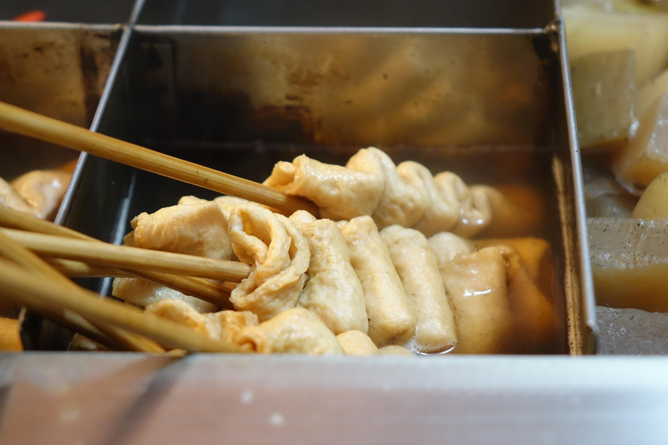 Close-up of Korean eomuk (fish cake skewers) simmering in hot broth at a street food stall, with folded pieces of fish cake threaded on wooden sticks and soaking in flavorful soup.