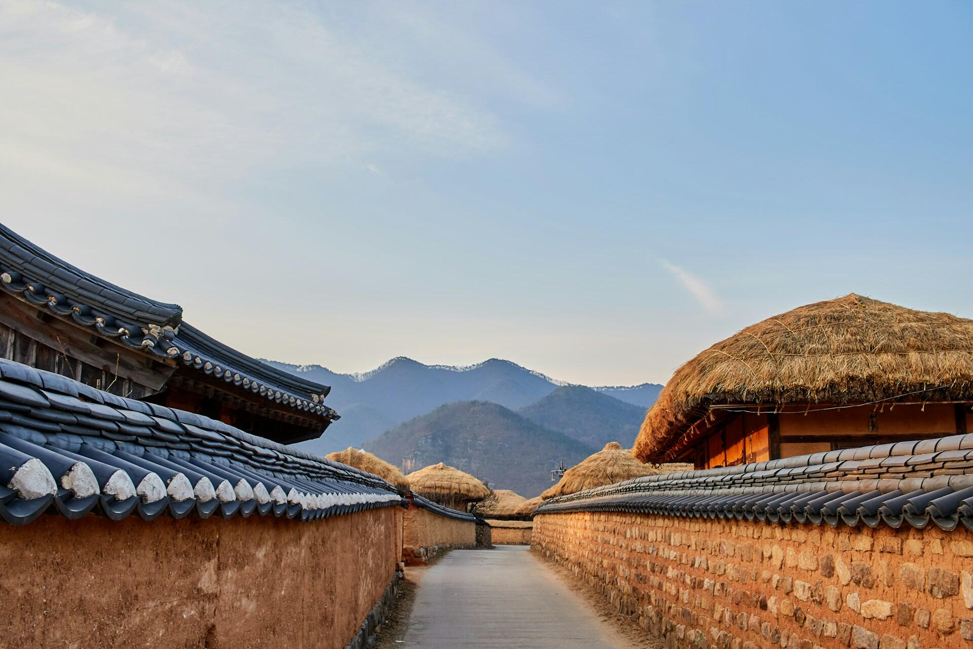 Traditional Korean village street with stone walls, thatched roofs, and mountains in the background under a clear sky.