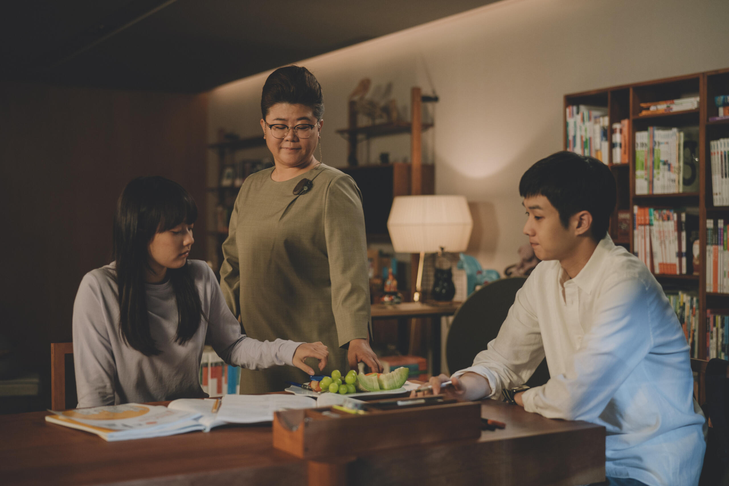 Three people in a cozy room, two seated studying with books, a third standing offering fruit on a plate.