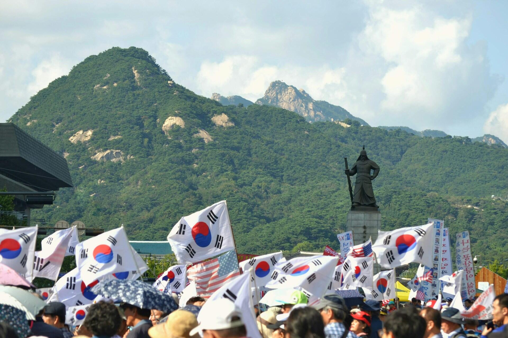 Crowd waving South Korean flags near a statue with green mountains and a partly cloudy sky in the background.