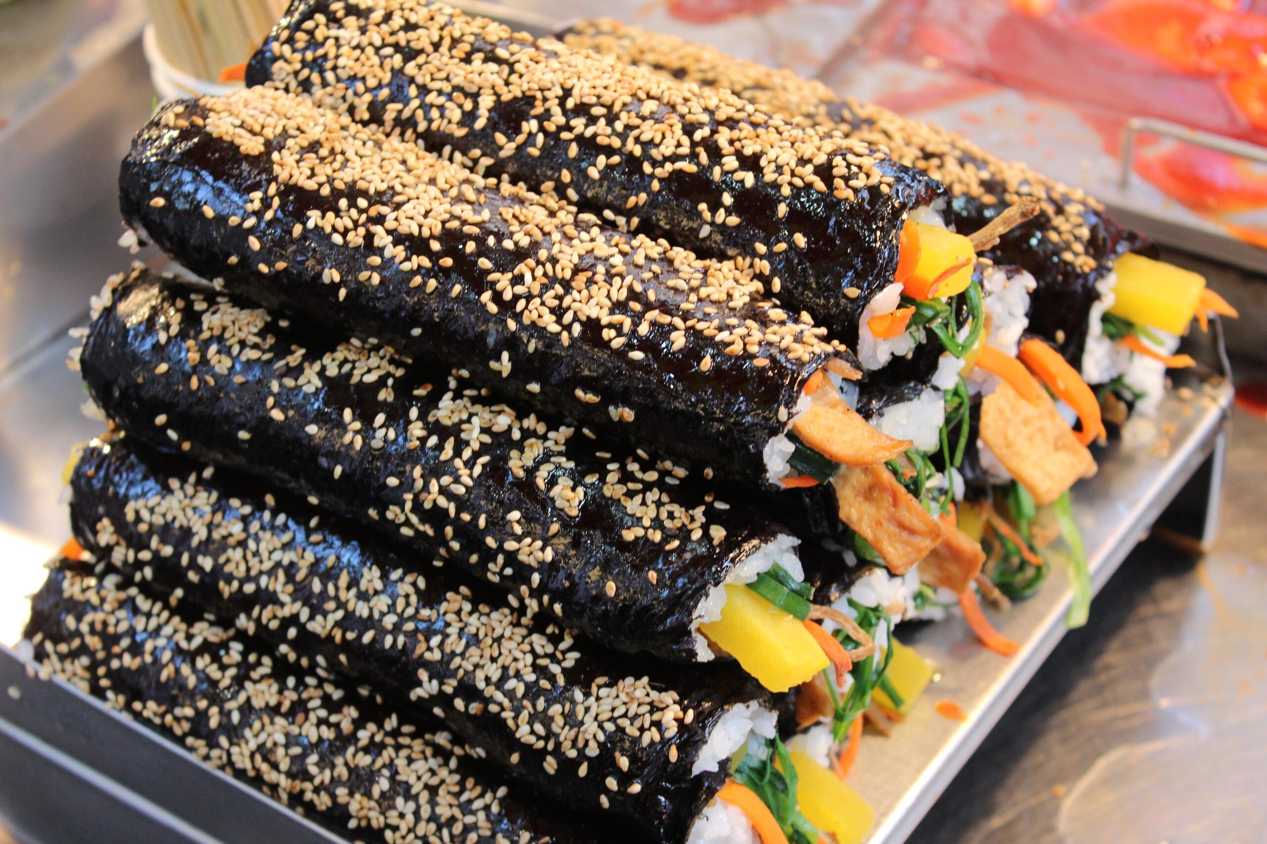 A stack of freshly made Korean gimbap rolls coated with sesame seeds, filled with rice, pickled radish, spinach, carrots, and fish cake, wrapped in shiny seaweed sheets, displayed at a street food stall.