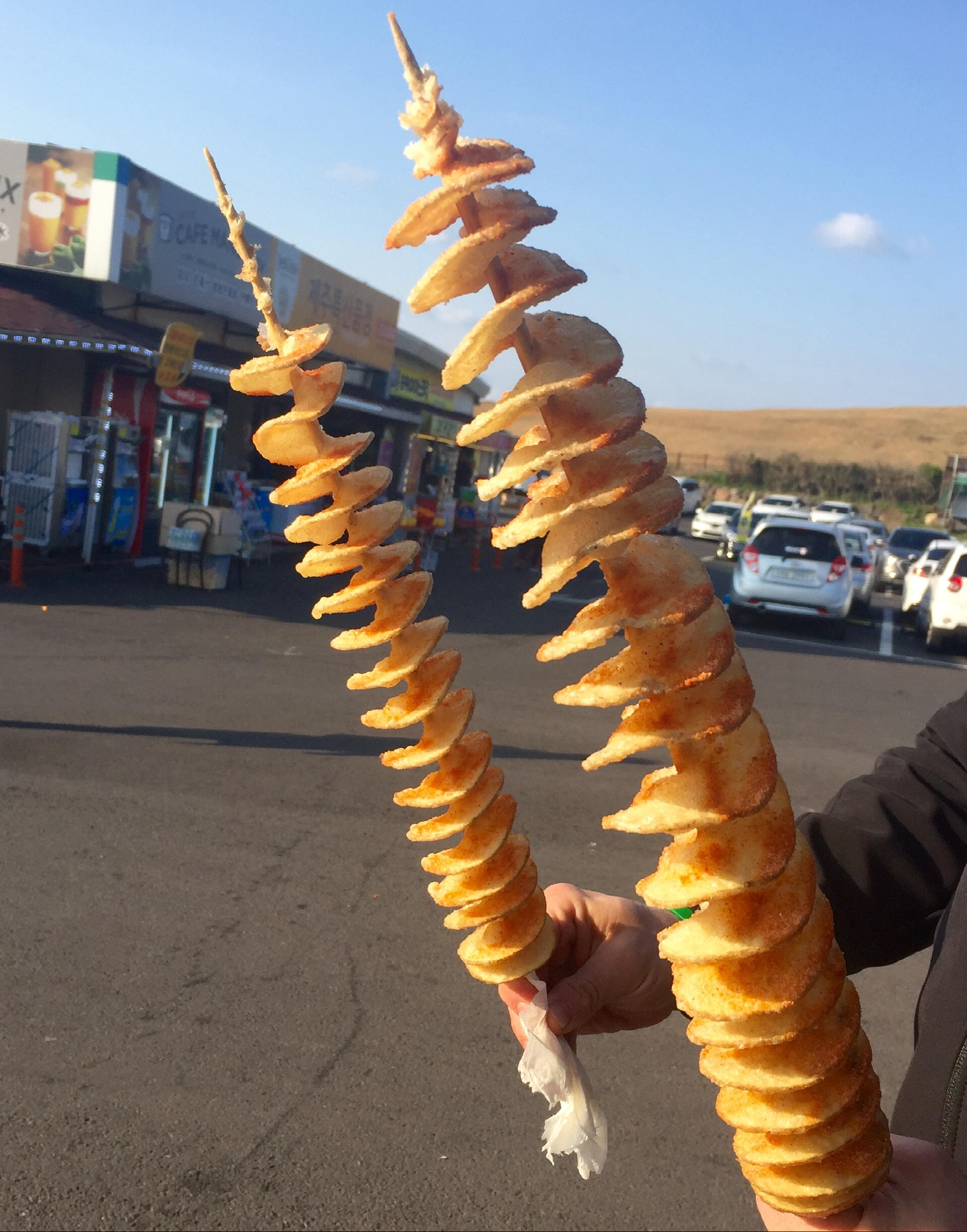 Two spiral-cut Korean tornado potatoes (hoeori gamja) on long skewers, deep-fried until golden and lightly dusted with seasoning, held outdoors near a street market with cars and shops in the background.
