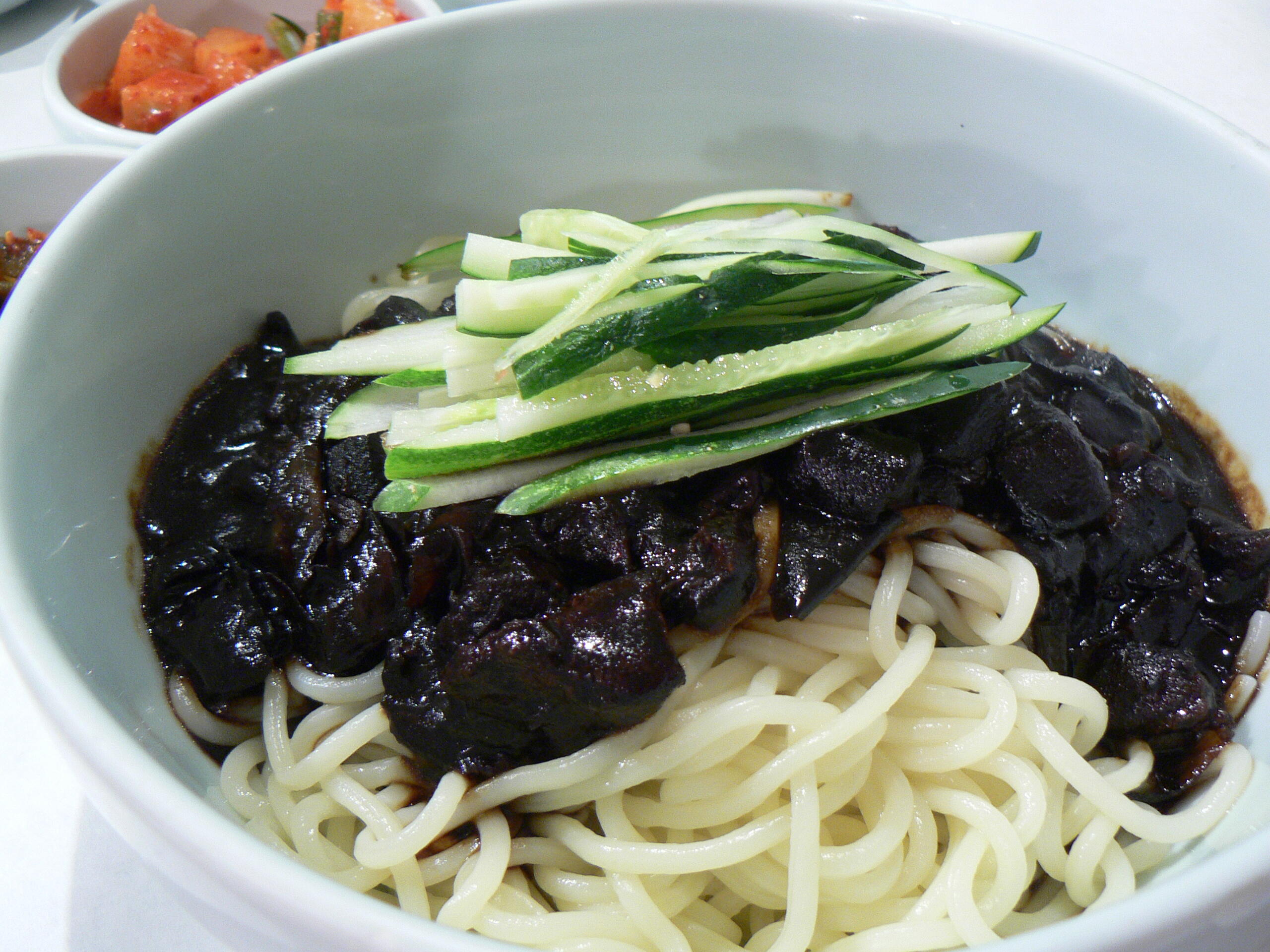 A bowl of Korean jajangmyeon noodles topped with rich black bean sauce, diced vegetables, and strips of fresh cucumber, served alongside small side dishes of kimchi.