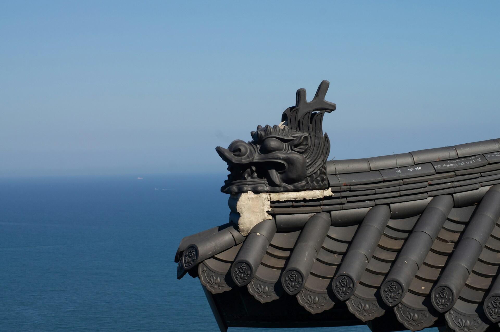 Traditional Asian-style roof corner with a black dragon sculpture overlooking the blue sea under a clear sky.