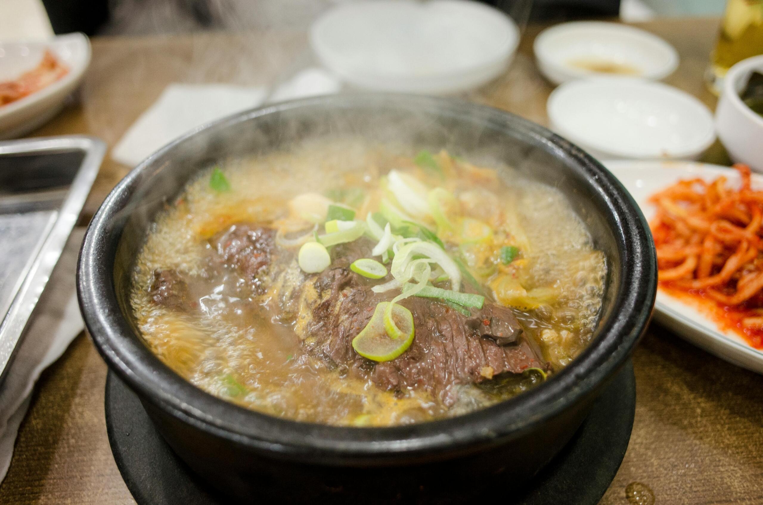A steaming bowl of Korean seolleongtang (ox bone soup) served in a black stone pot, topped with sliced green onions and tender beef, with side dishes like kimchi visible in the background.