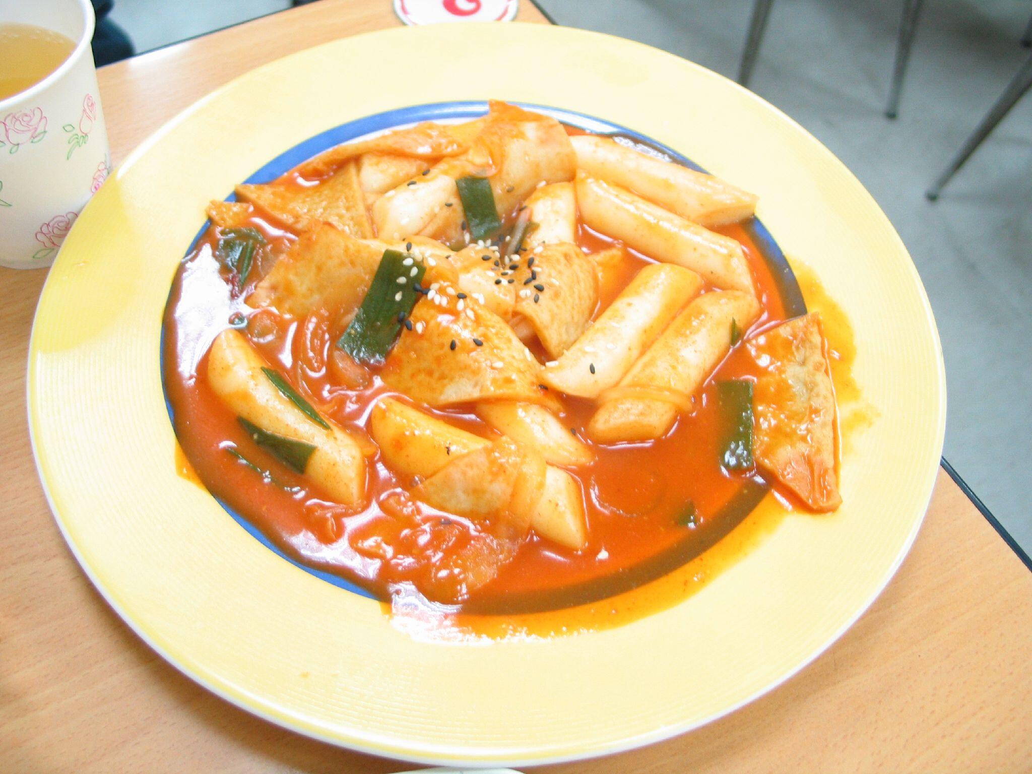 A plate of tteokbokki, a popular Korean street food made of chewy rice cakes simmered in a bright red gochujang (chili paste) sauce, garnished with sesame seeds and sliced vegetables, served on a yellow-rimmed plate.