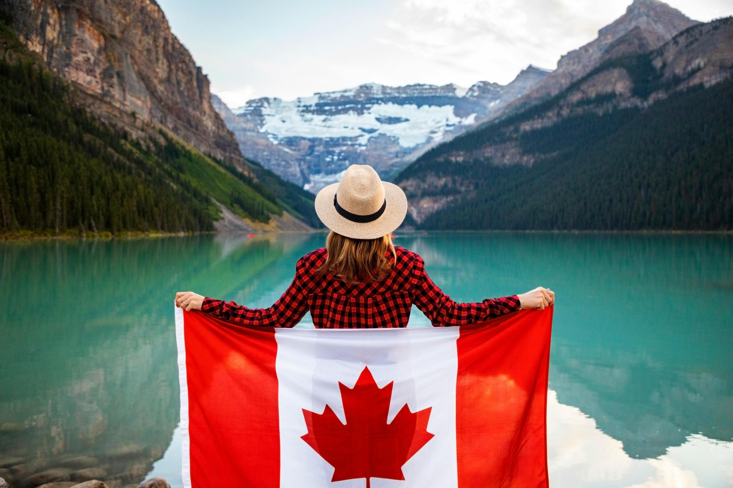 a woman stands in front of a scenic lake holding a Canada flag.