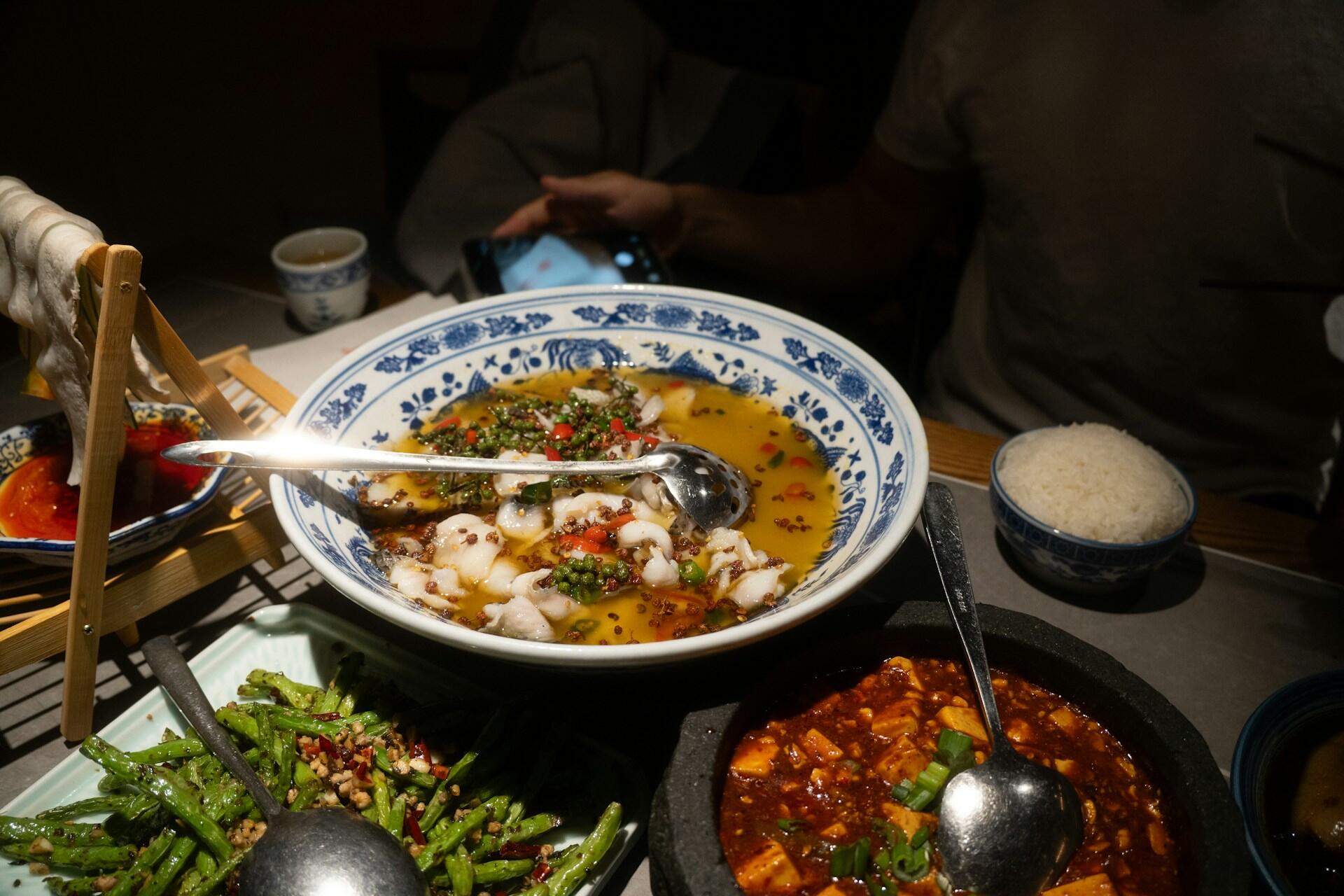Table with Chinese dishes including mapo tofu, fish in chili oil, stir-fried green beans, a bowl of rice, and bamboo rack with hanging noodles.
