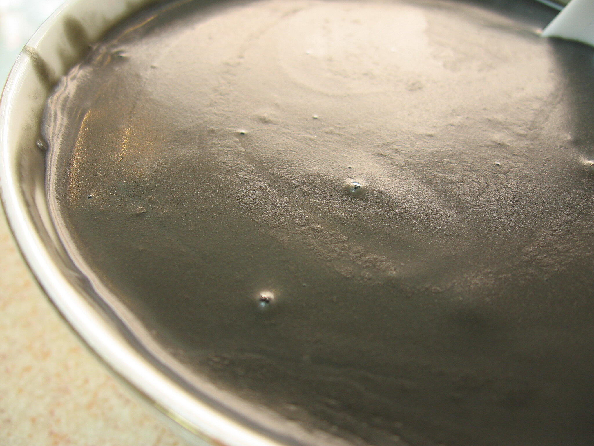 Close-up of a smooth, dark gray mixture in a metal bowl, with small air bubbles on the surface glistening in soft light.