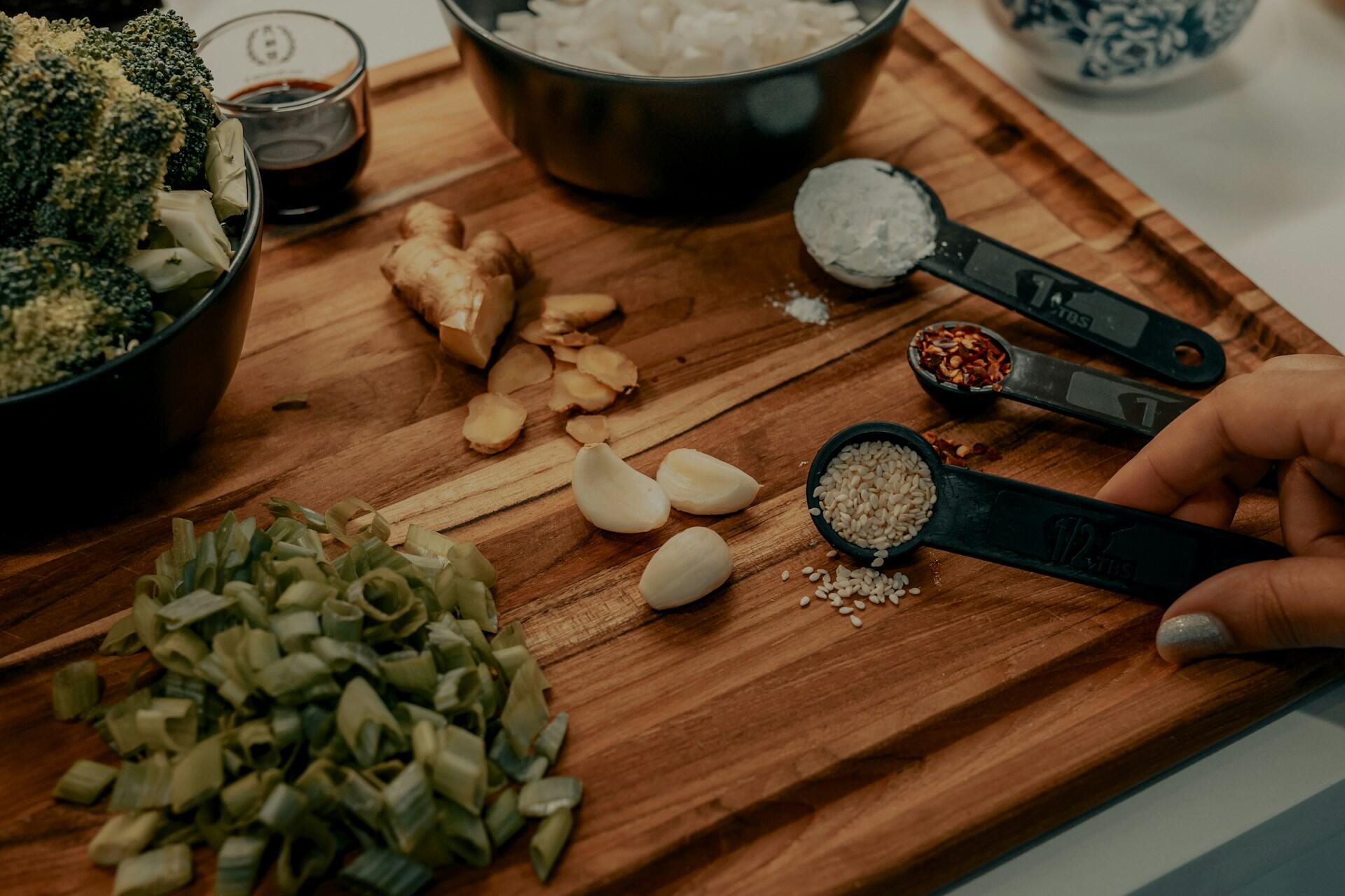Hand holding a measuring spoon with sesame seeds on a wooden board surrounded by garlic, ginger, broccoli, and spices.