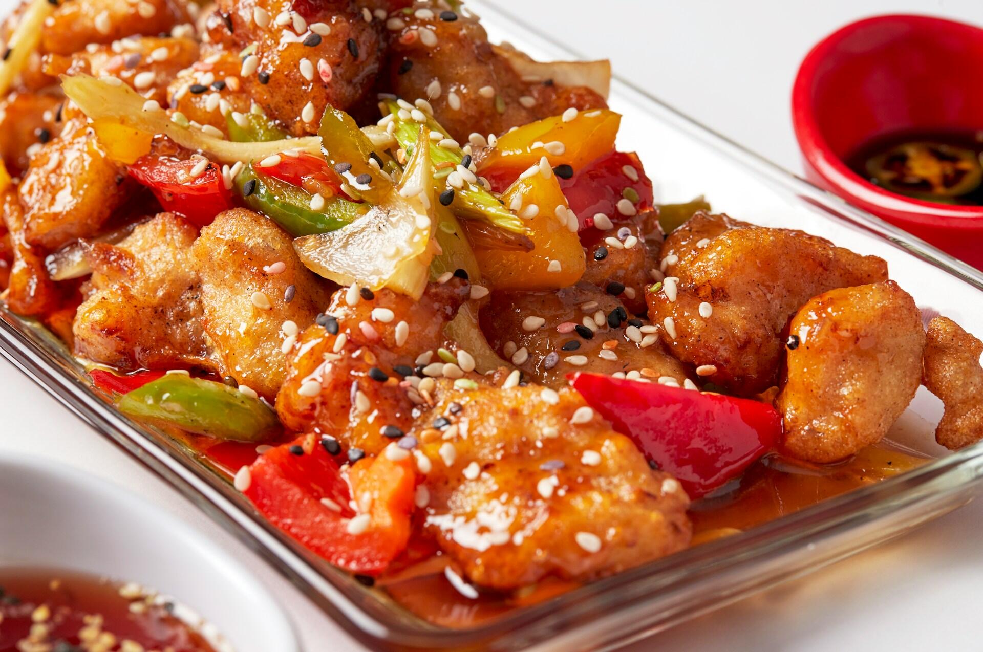 Glazed fried chicken pieces with bell peppers, onions, and sesame seeds in a glass dish, with red and white bowls of dipping sauce.