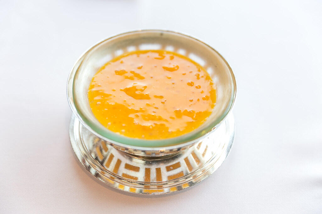 A silver bowl filled with bright orange dessert, glistening with a smooth, syrupy texture, resting on a decorative plate.