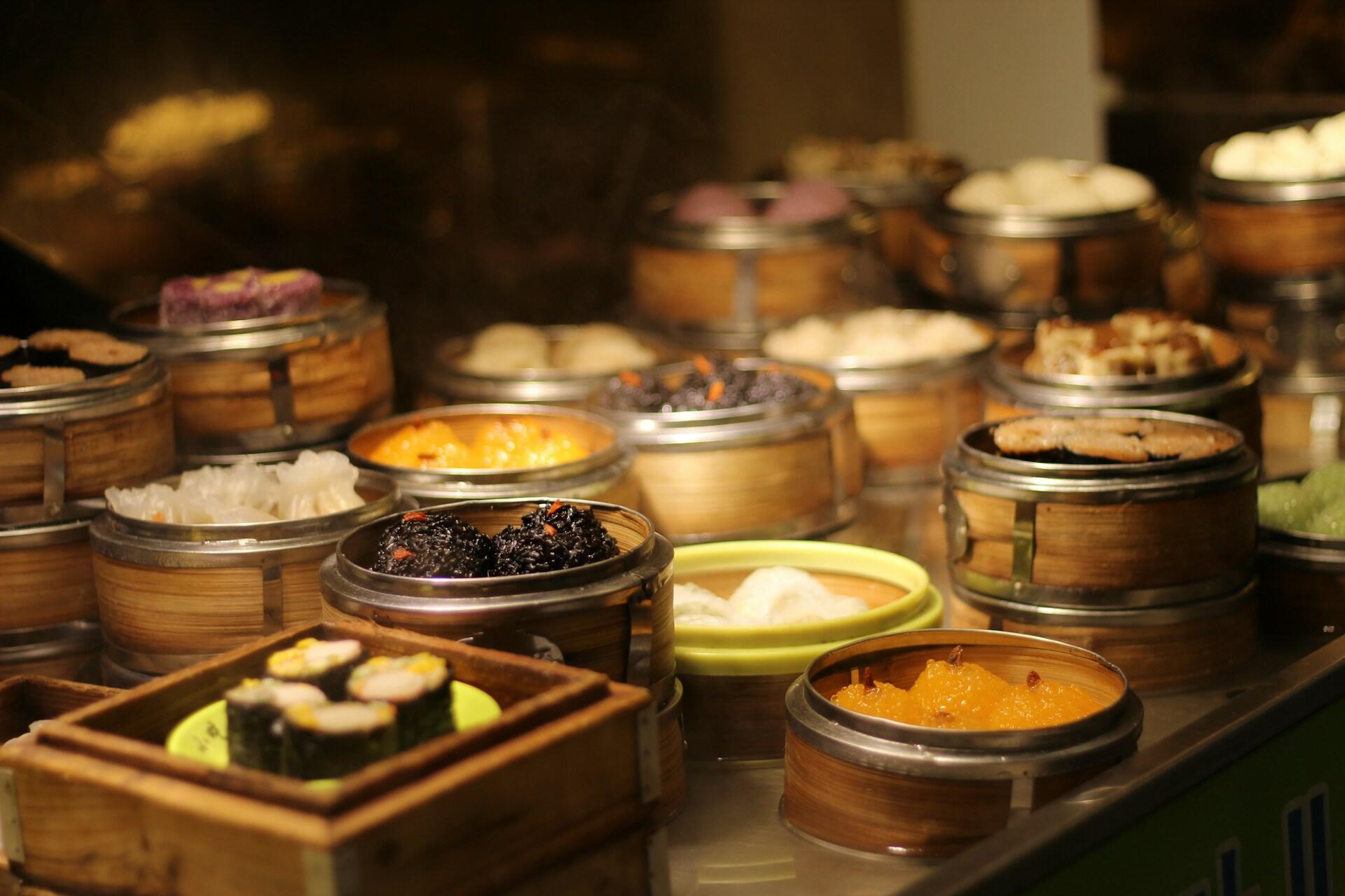 Assorted dim sum dishes in bamboo steamers displayed on a counter, featuring colorful dumplings and sushi rolls.