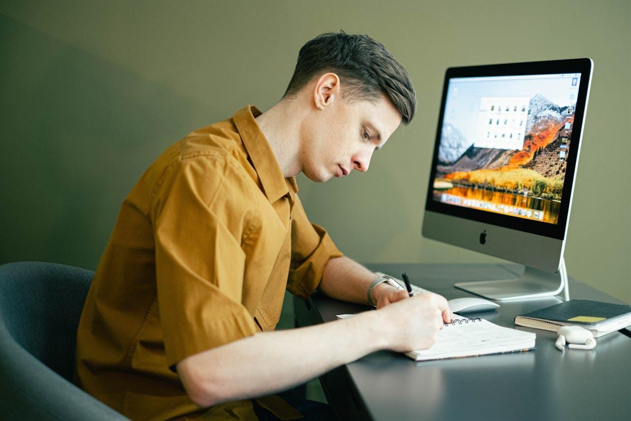A male studying for the NCLEX with a computer and a notebook. 