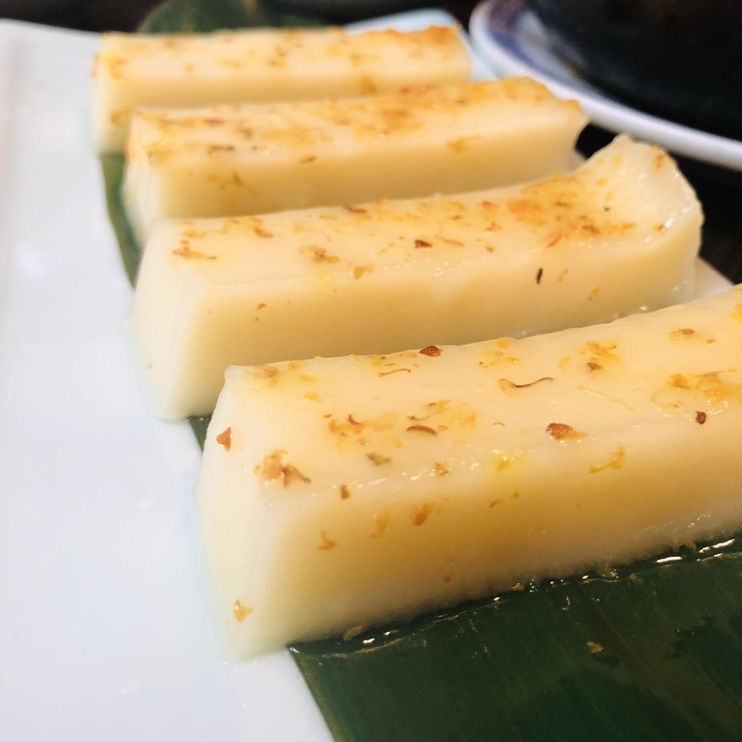 Four rectangular slices of creamy coconut dessert topped with golden-brown flakes, served on a banana leaf.