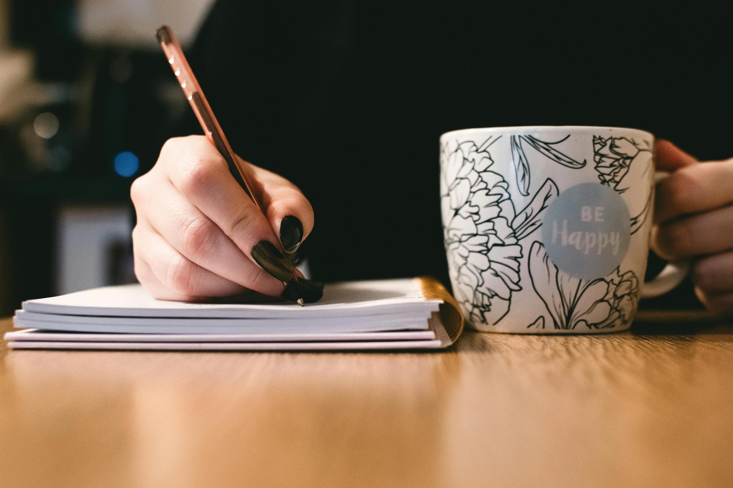a person writes in a notebook while holding a. mug.