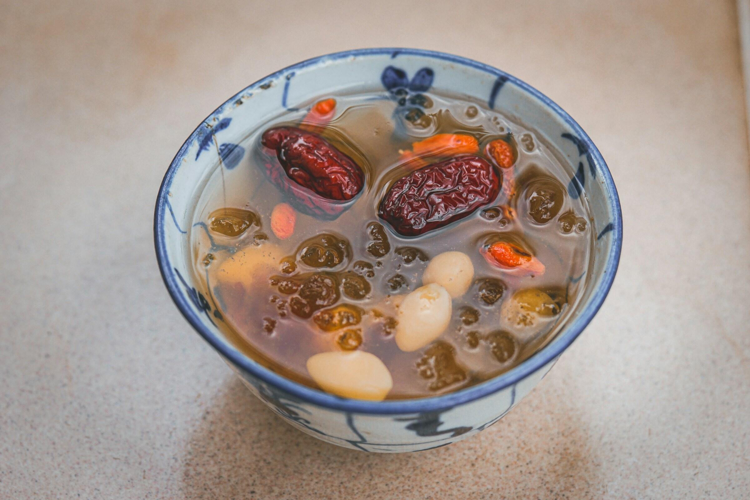 A bowl of herbal soup with dates, goji berries, and white nuts, resting on a smooth surface.