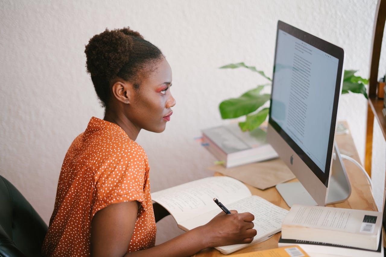A lady taking notes at her desk with a notepad as she reads from a computer.