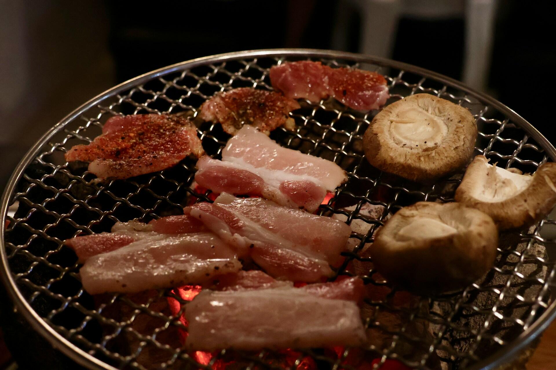 Slices of meat and whole mushrooms grilling on a round wire mesh over hot charcoal.