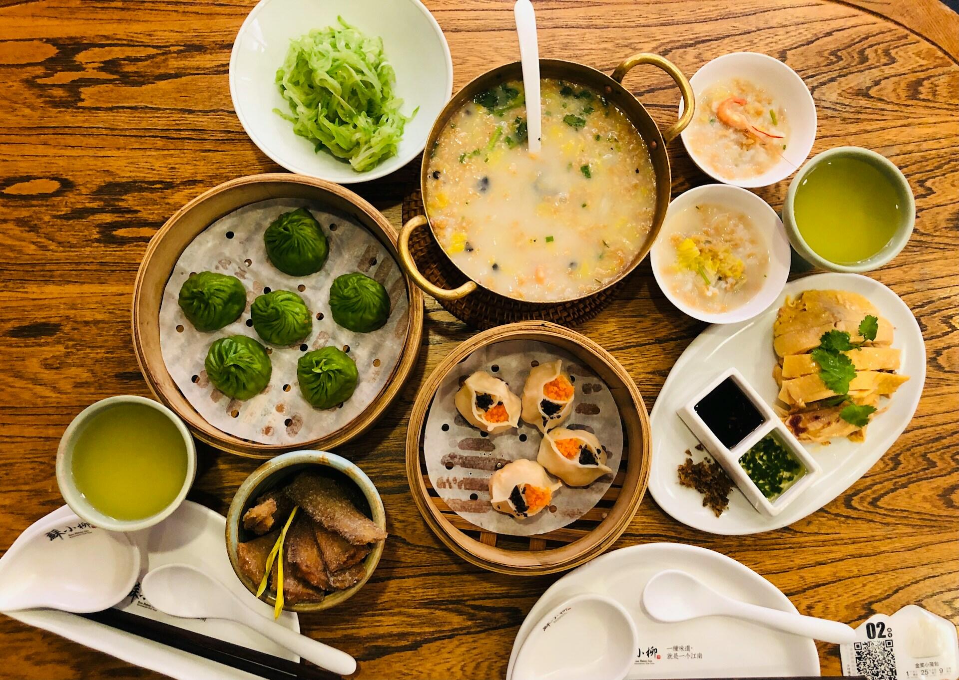 Top-down view of a wooden table with various Asian dishes including green dumplings, soup, chicken, dumplings with fish roe, green tea, and vegetables.