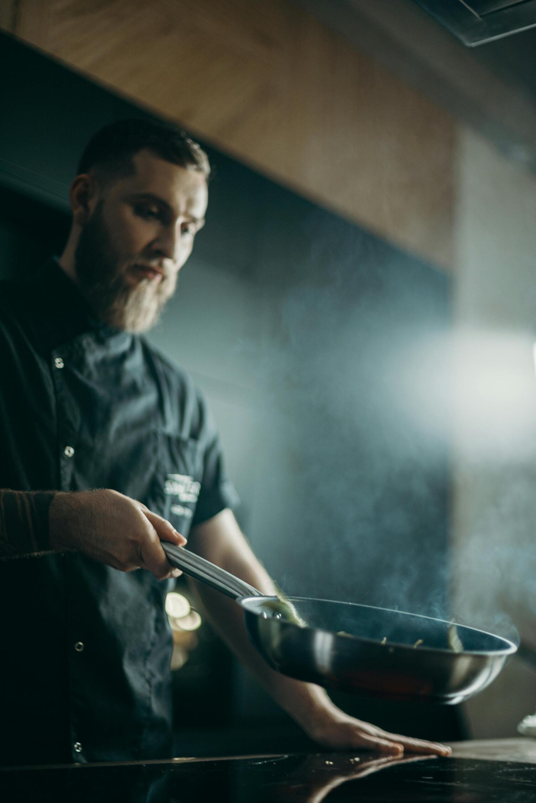 A bearded man stands holding a frying pan confidently in front of him, smiling slightly as he poses in a kitchen setting.