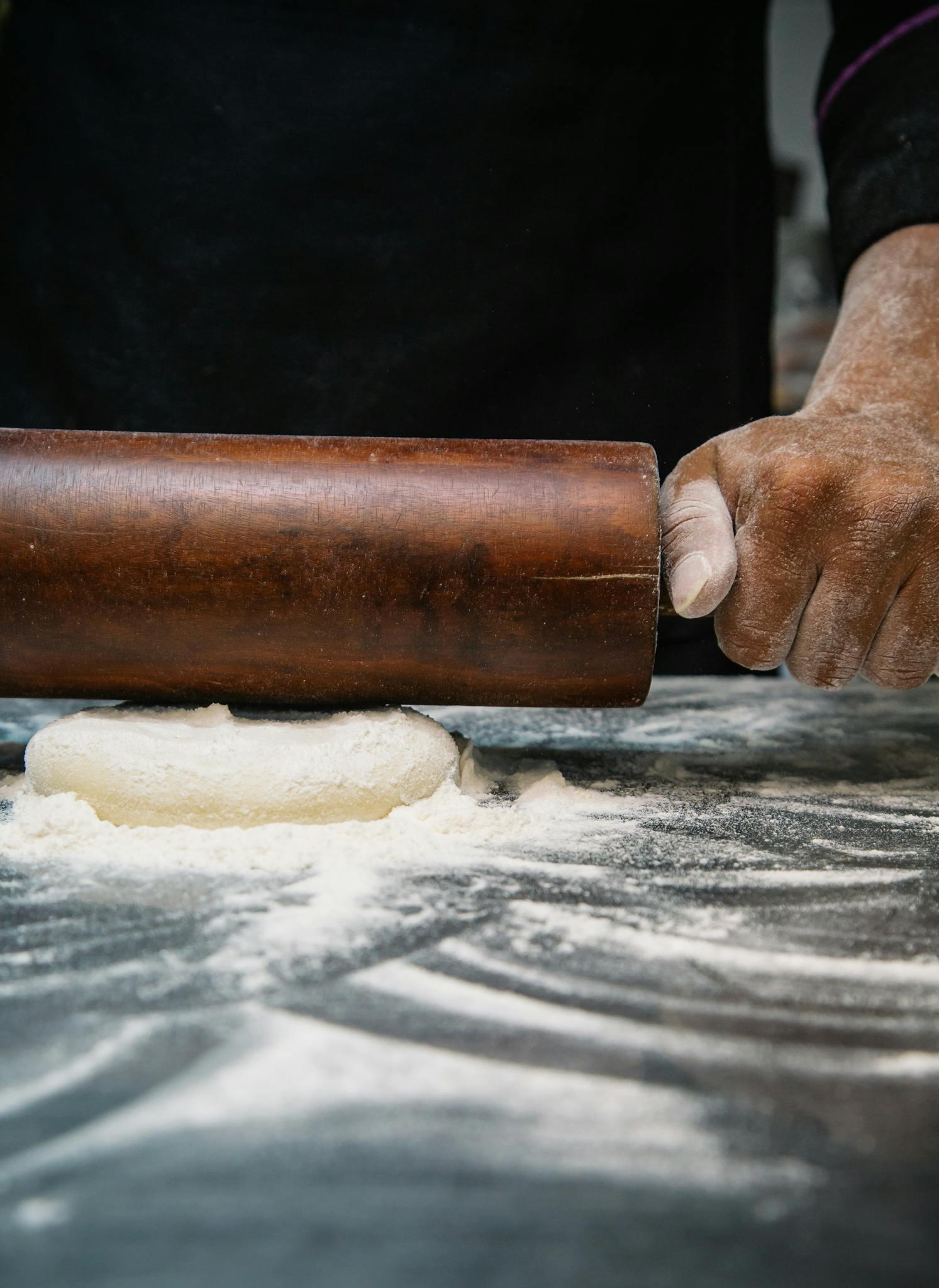 A pastry chef rolls out a sheet of dough on a floured work surface, using a wooden rolling pin with focused, precise movements.