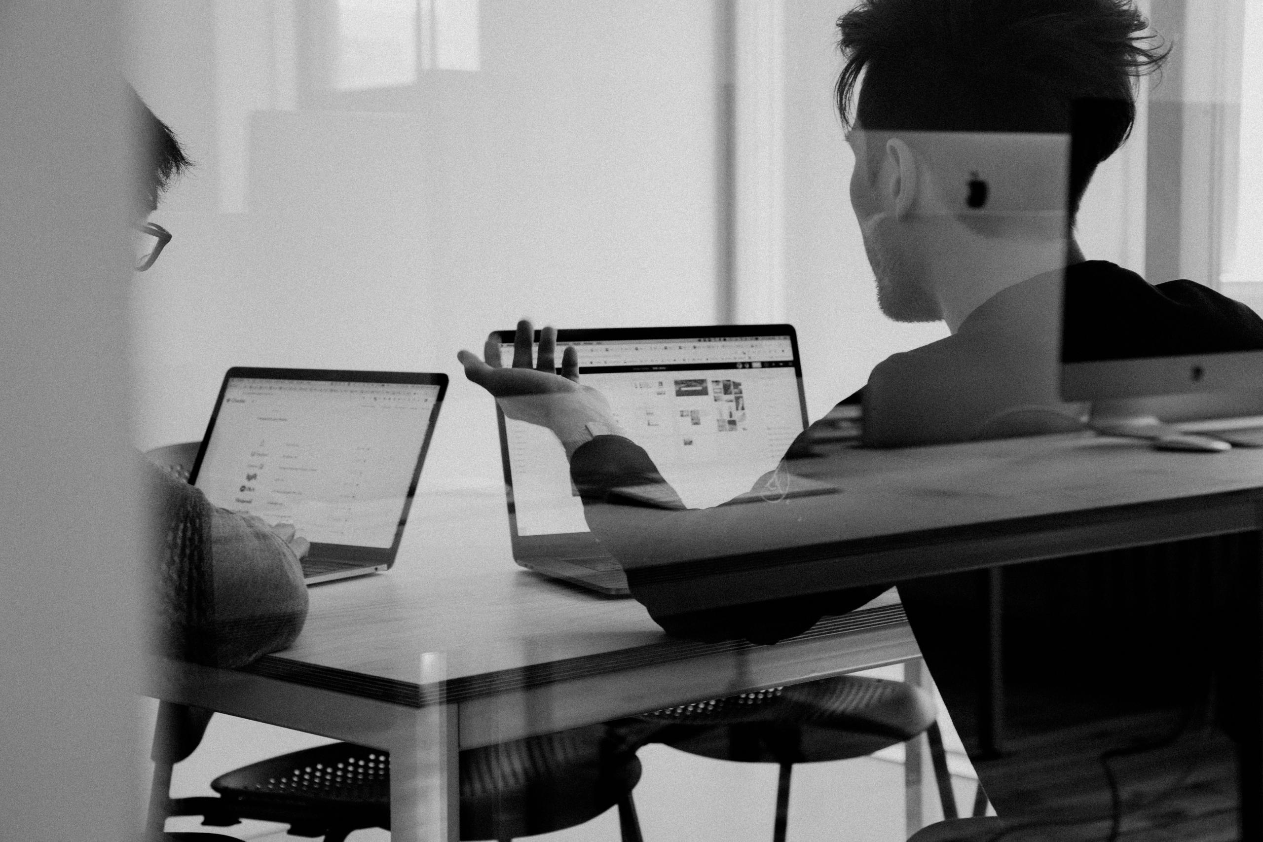 A Graphic designer talking to a client. They are sitting at a table with their computers. The Graphic designer is a a man and he's gesturing with his arm.