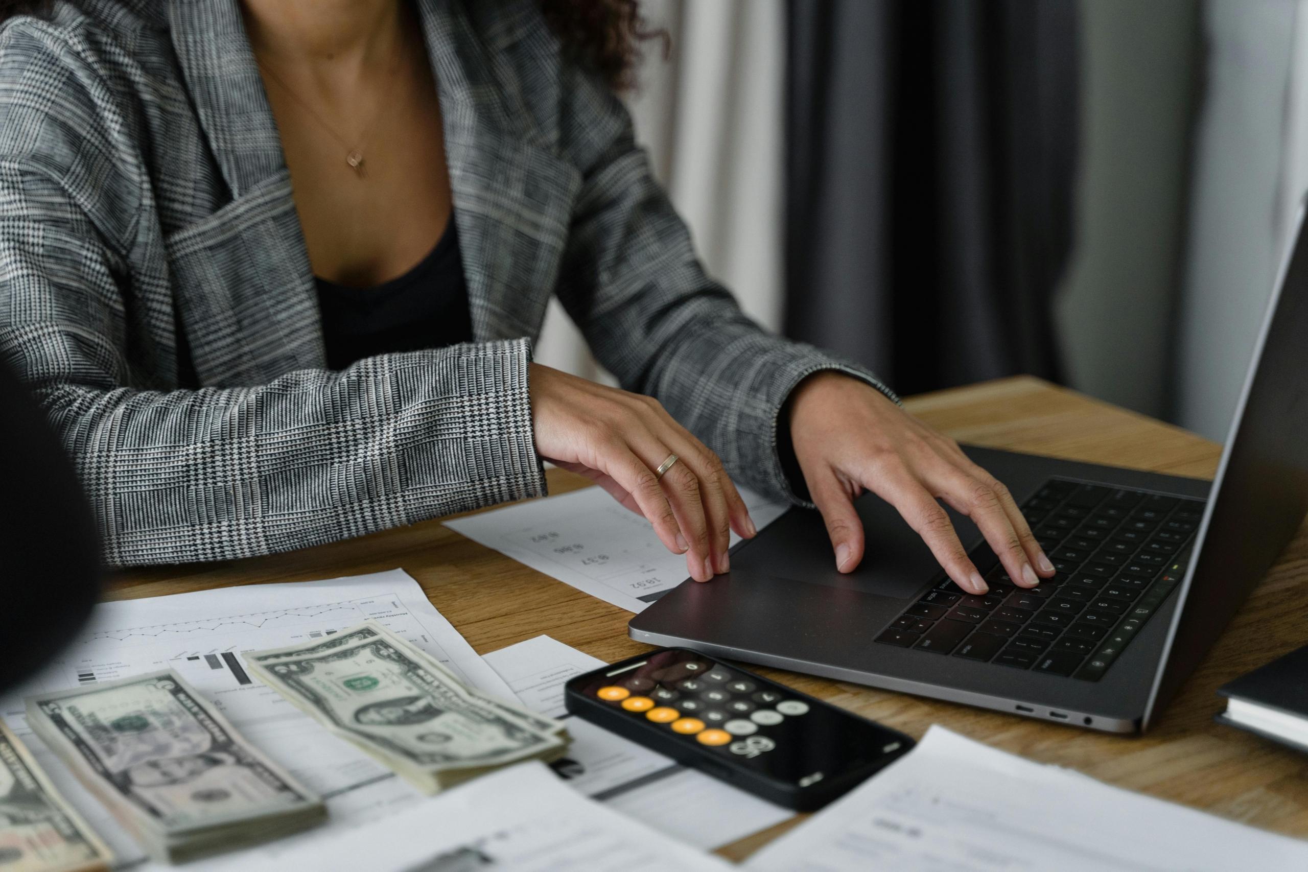 A businesswoman works on a laptop. A calculator, documents, and cash are on the desk.