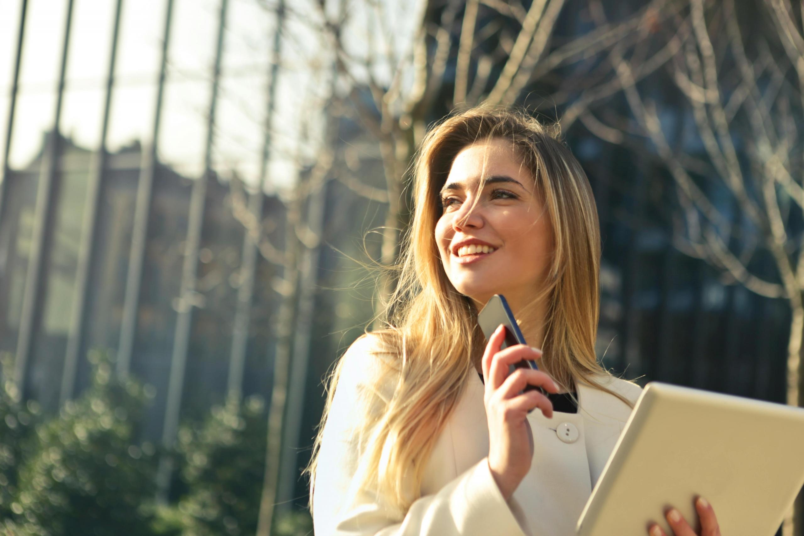 A professionally dressed woman stands outside on a sunny day. She holds a tablet and a smart phone.