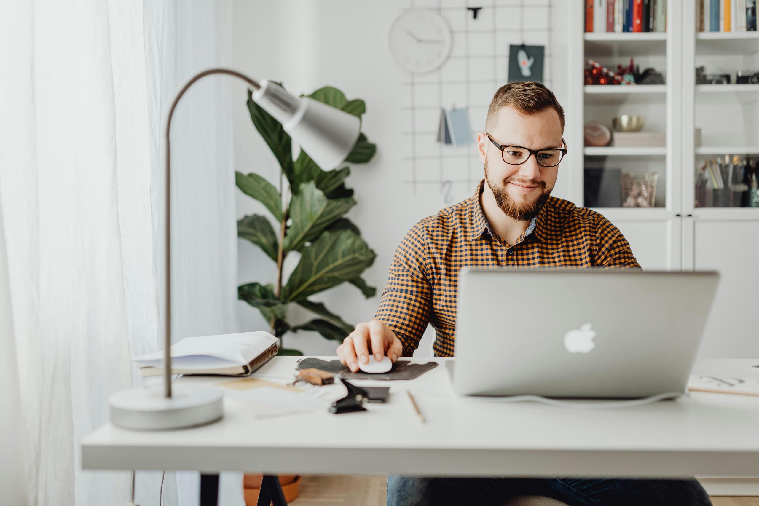 A smiling man in a plaid shirt works at a laptop.