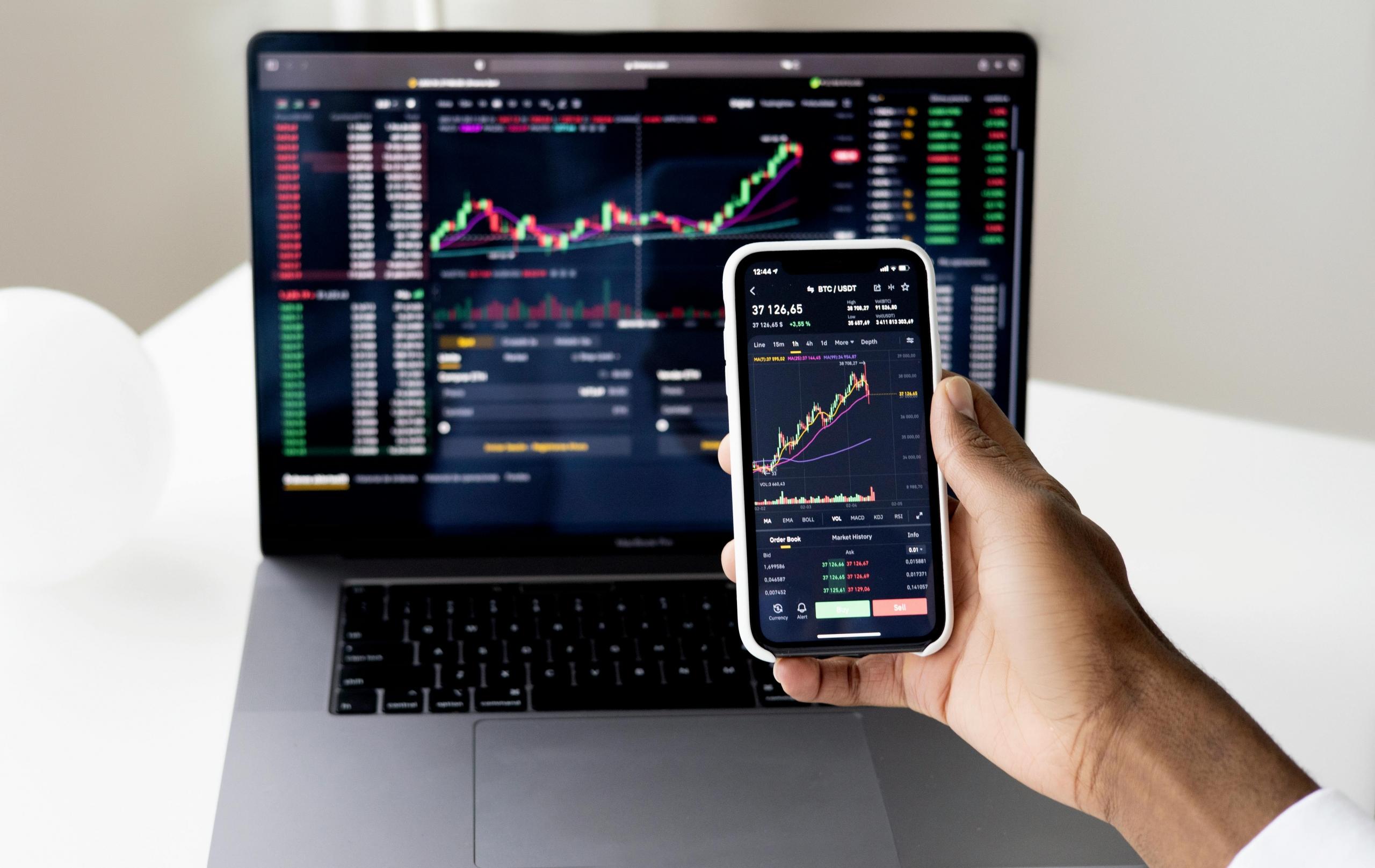 A person holds a smartphone up with information related to the stock market. A laptop with similar data and charts is in the background.