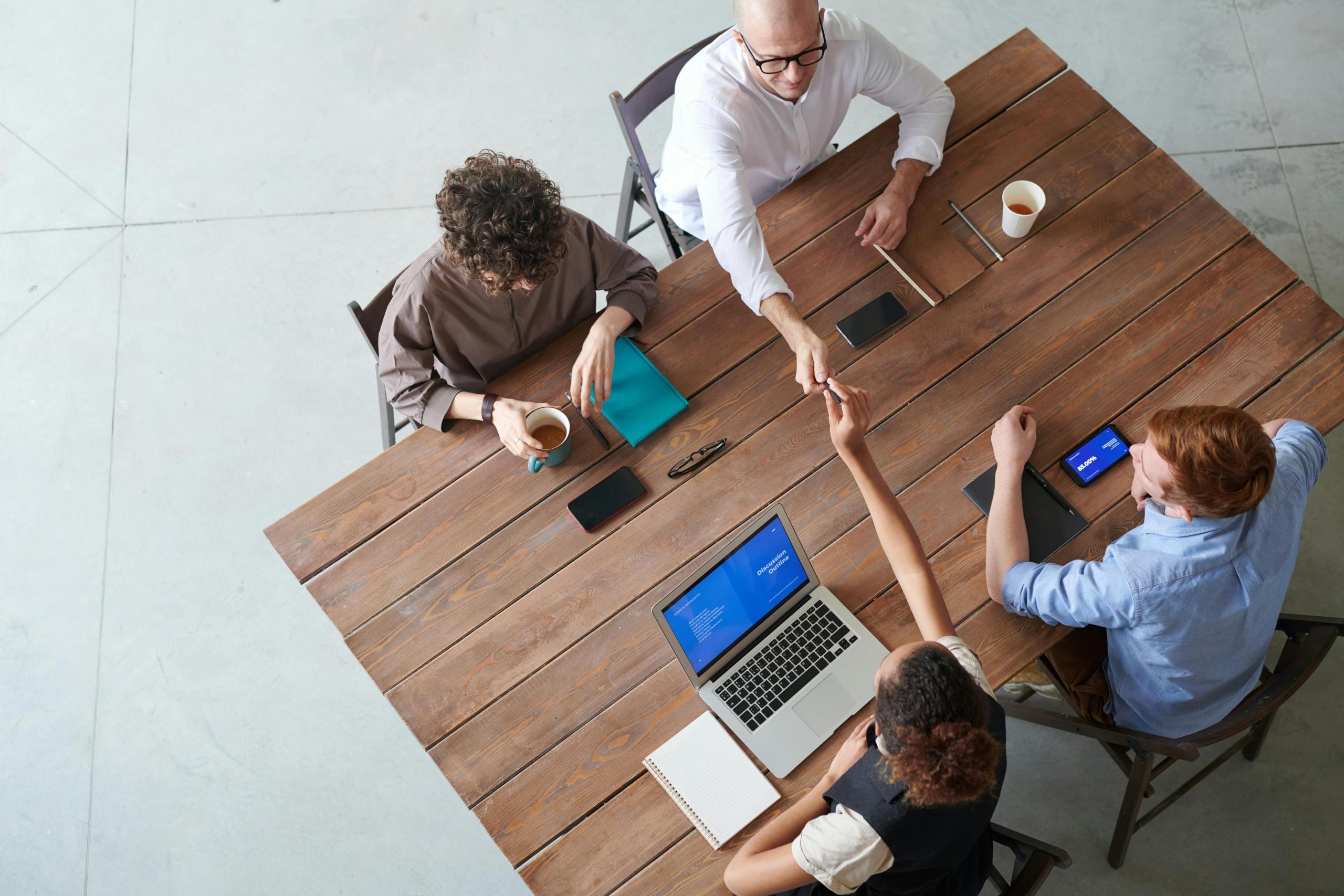 Four professionally dressed individuals are engaged in a meeting. They drink coffee and each have mobile devices.
