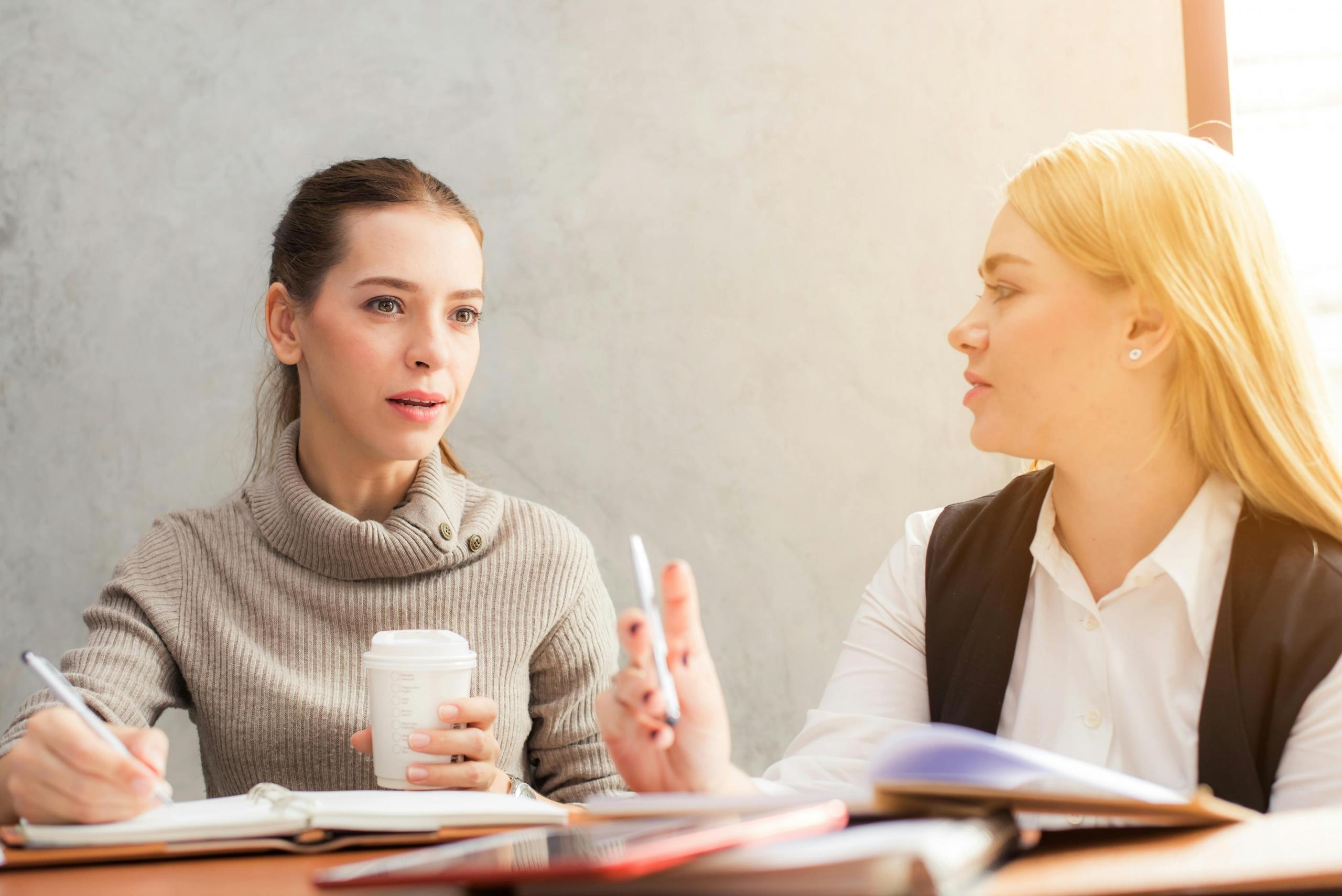 A woman holding a coffee works with another woman. There is a stack of notebooks and a tablet on the table in front of them.