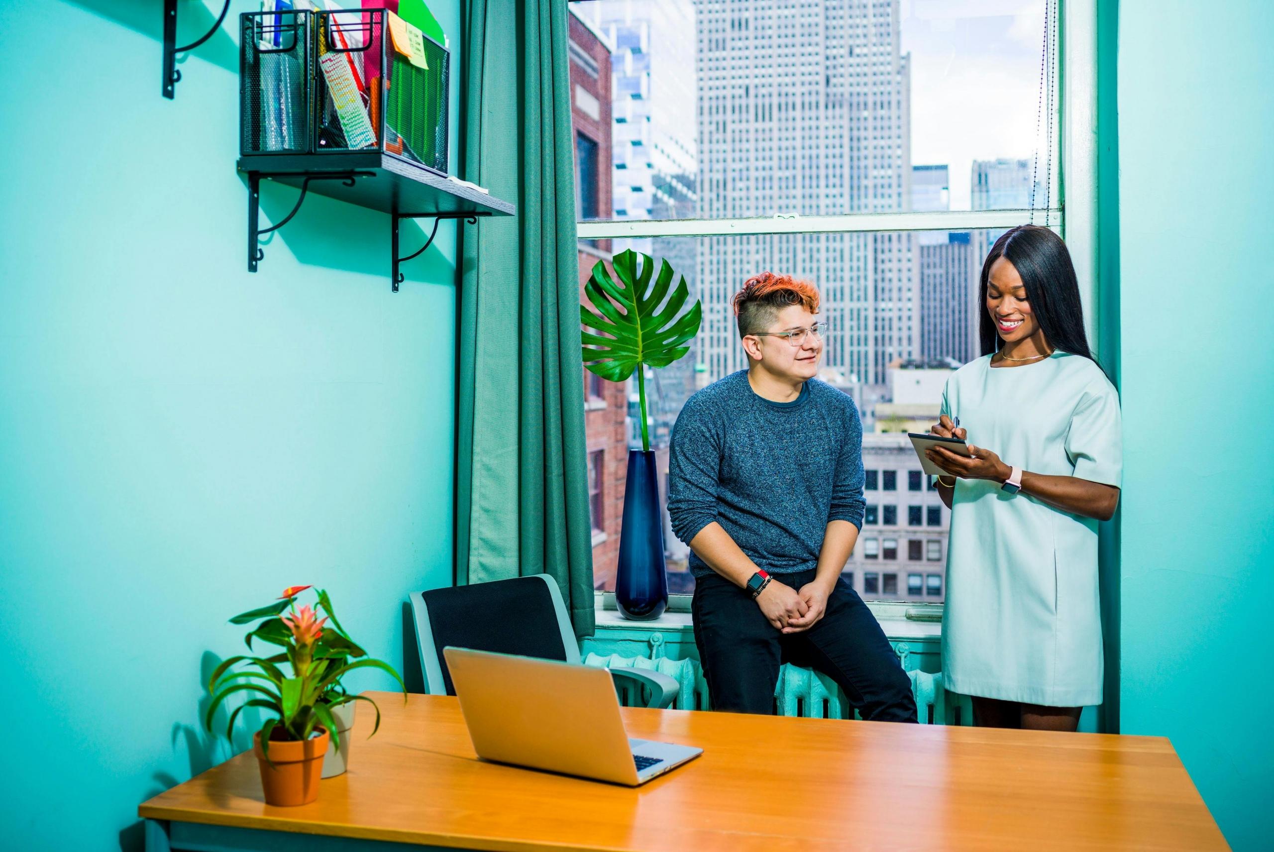 two professionals stand in a brightly painted turquoise office in front of a desk with a laptop.