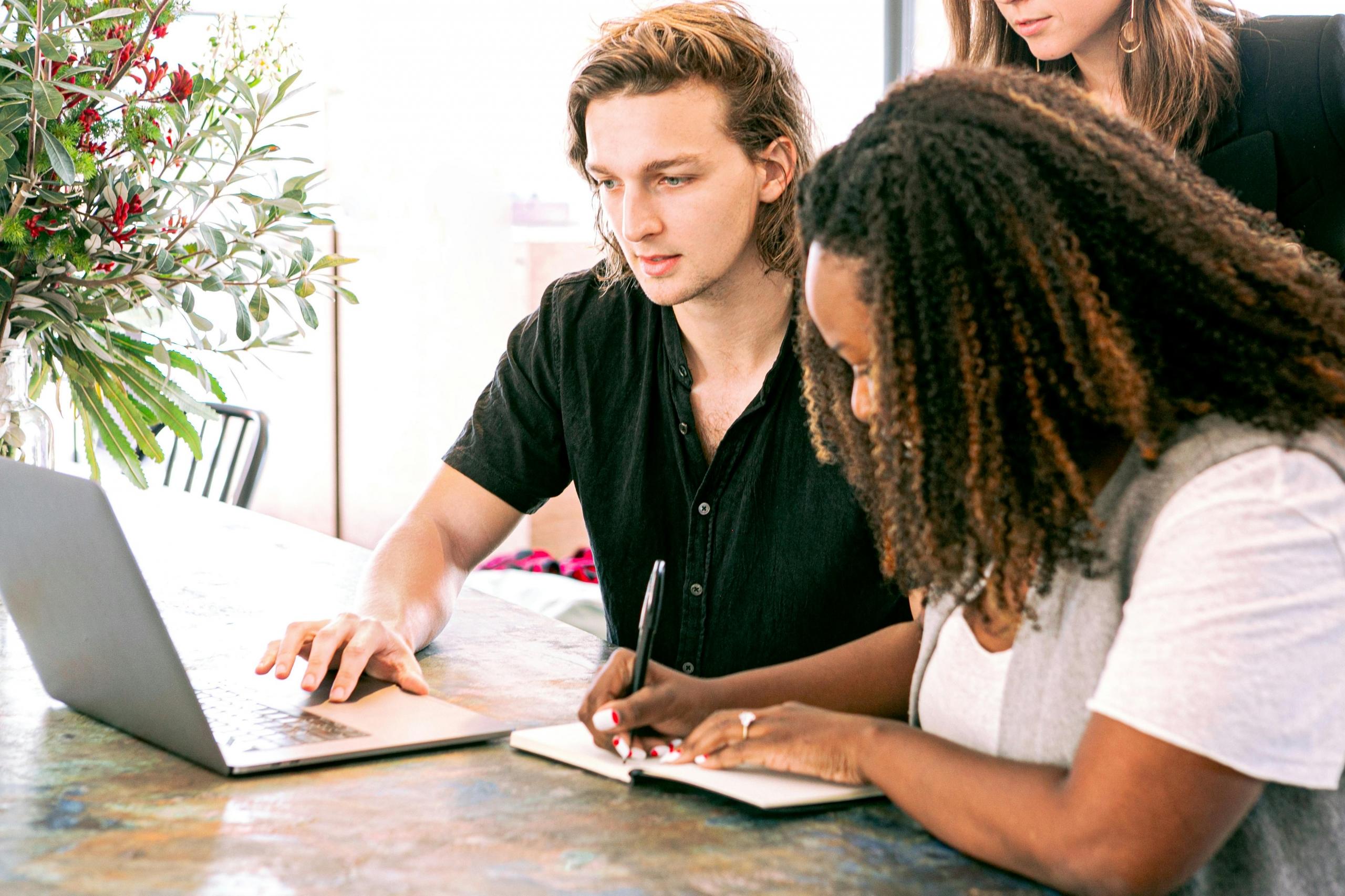 a man works on a laptop beside a woman. The woman is writing notes her in a journal.