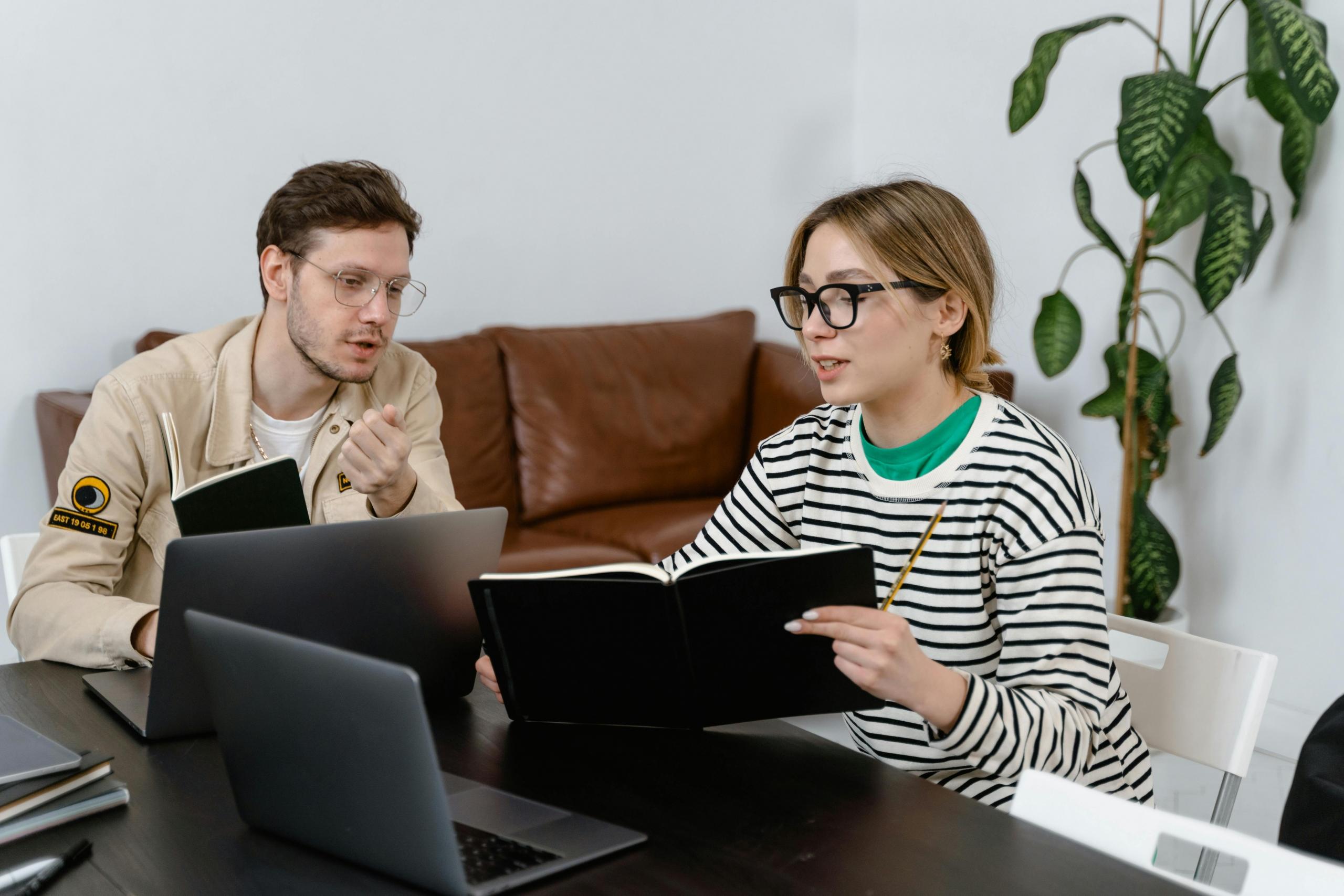 A man works in front of a laptop with a teenager. The teenager holds a notebook open.