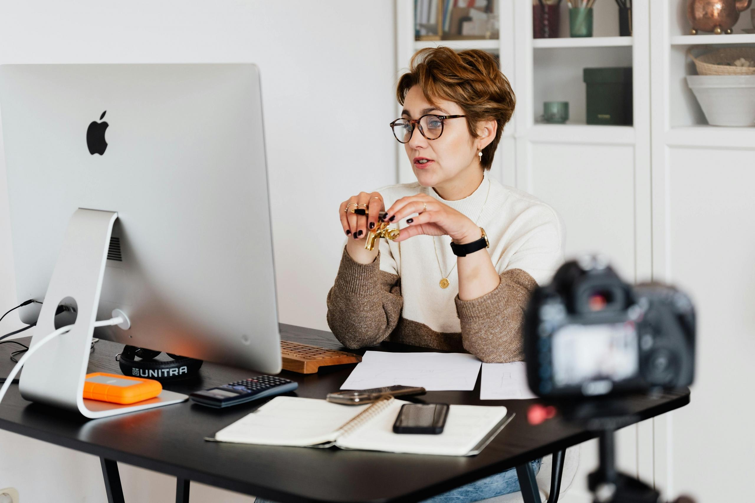 A woman with short hair and glasses looks into a large computer moitor. There are papers and a cell phone on her desk.