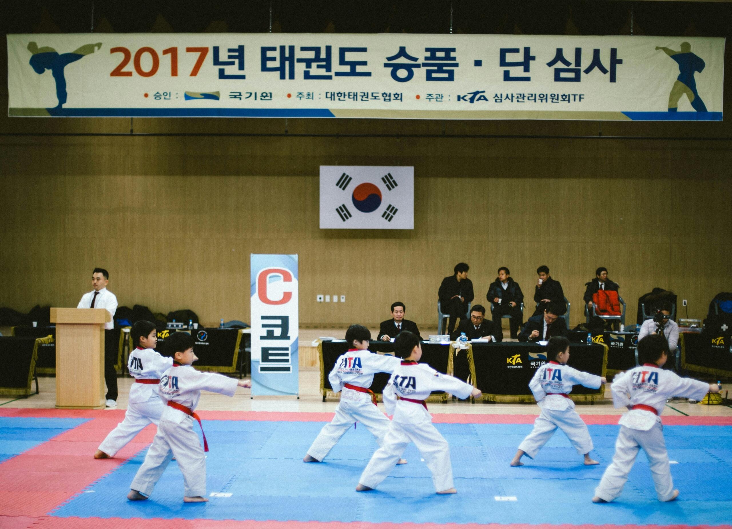 Young taekwondo practitioners perform in a competition setting, with judges and spectators observing in the background.