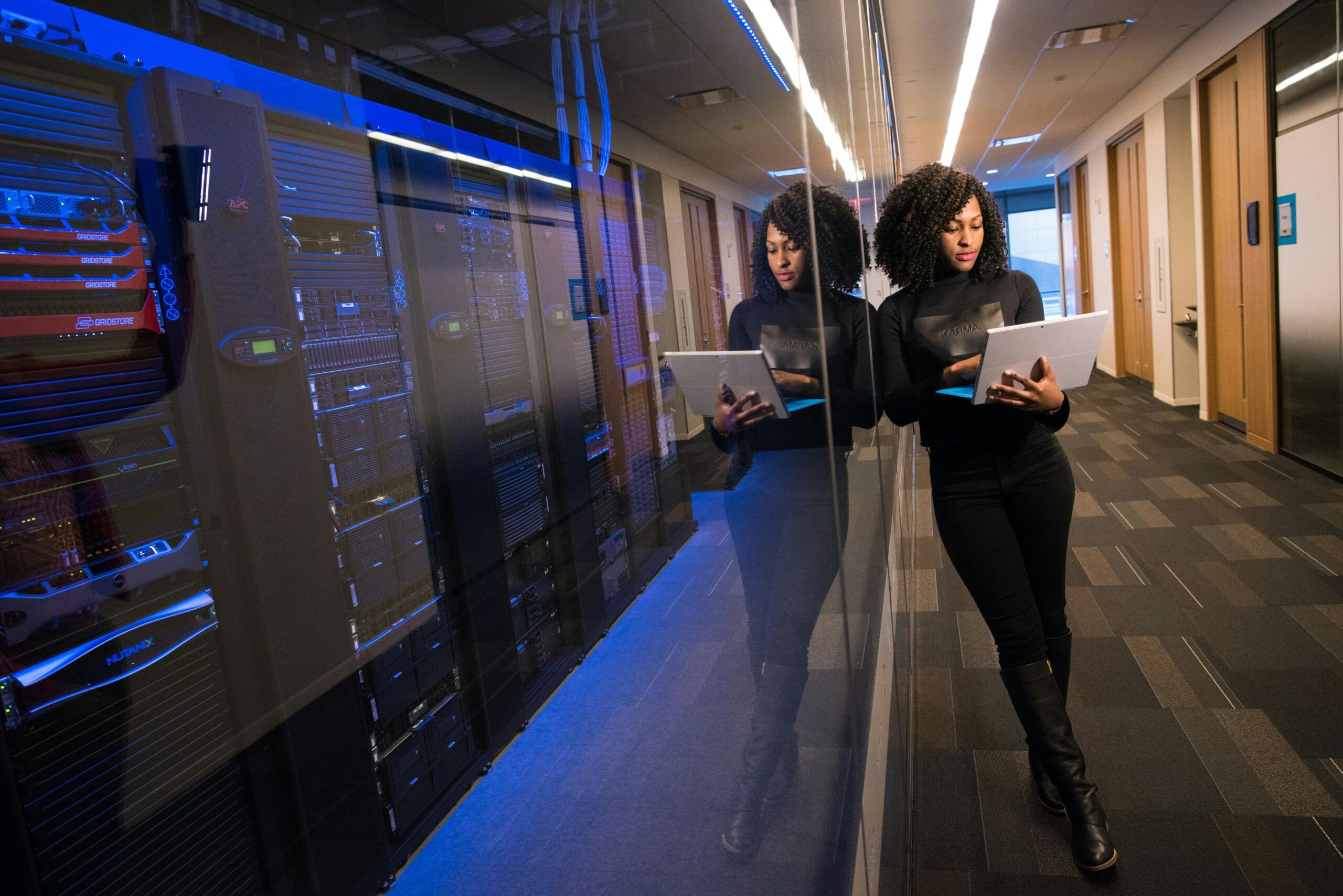 A woman stands in a hallway next to server racks, working on a laptop, with her reflection visible in the glass wall.
