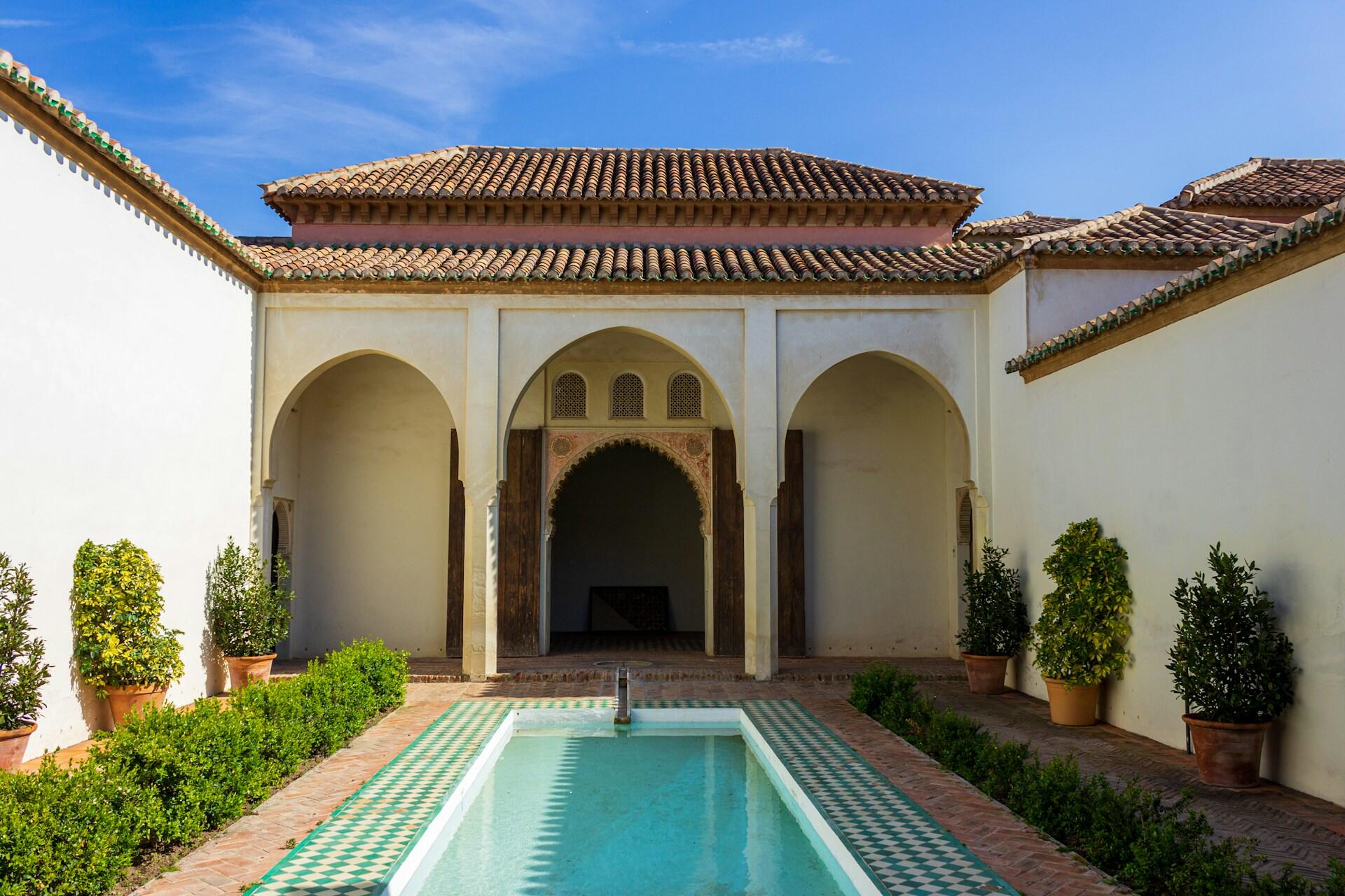 A serene courtyard featuring a reflecting pool, potted plants, and arched doorways under a clear blue sky. Traditional Moroccan architecture.