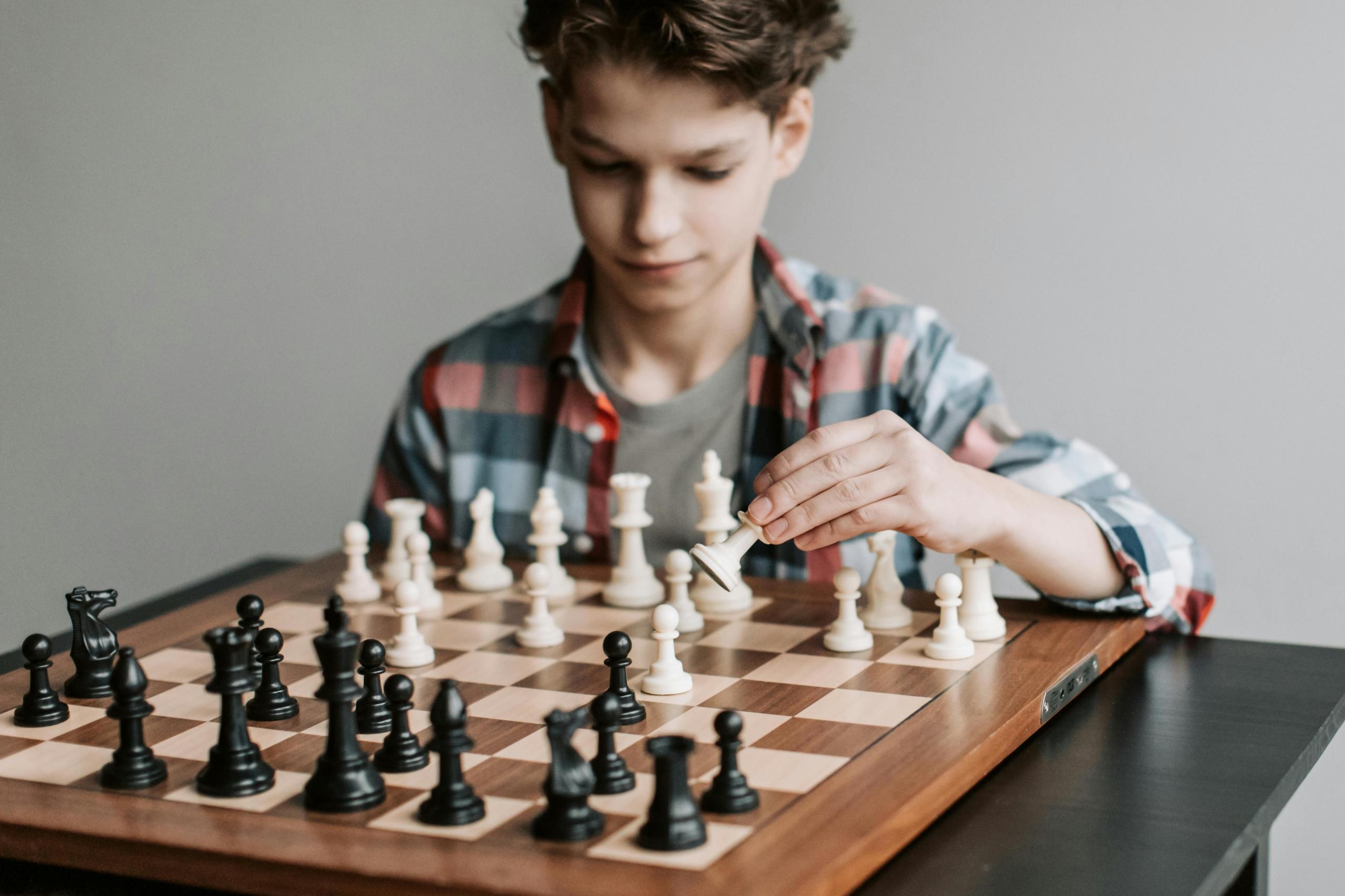 A boy about 12 years old plays chess. He holds a white piece. He is wearing a plaid shirt.